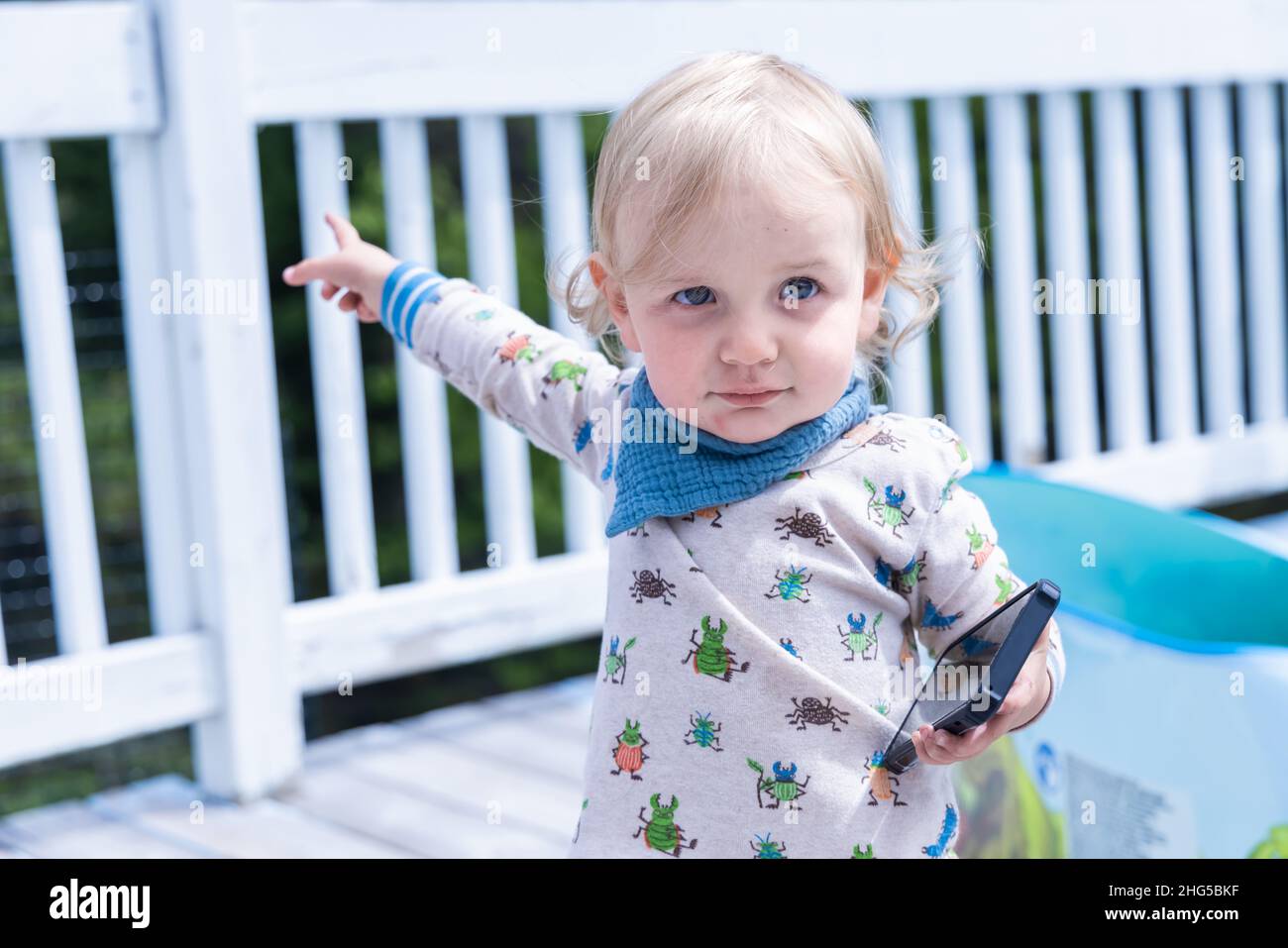 Portrait en gros plan d'un garçon de 3 ans, avec un teint blanc pâle et un joli petit cultivateur tenant un téléphone mobile dans l'arrière-cour pointant derrière. Banque D'Images