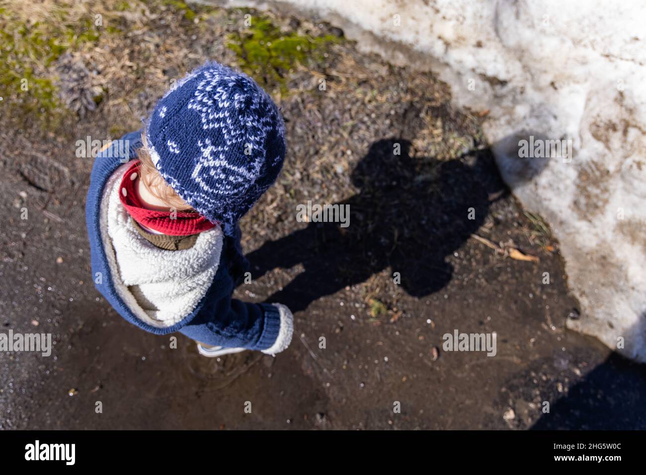 Photo en grand angle d'un enfant de 3 ans jouant à l'extérieur en hiver.Jeune garçon regardant l'ombre vue d'en haut portant des vêtements d'hiver et un chapeau. Banque D'Images