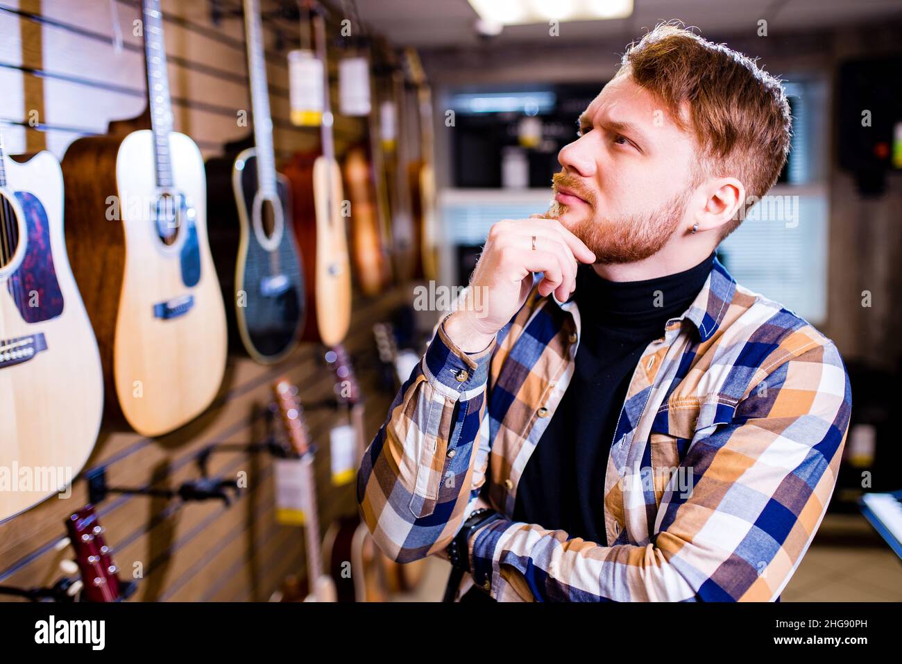 homme beau barbu à poil rouge en tissu écossais brun décontracté choisir une guitare dans un magasin de musique Banque D'Images