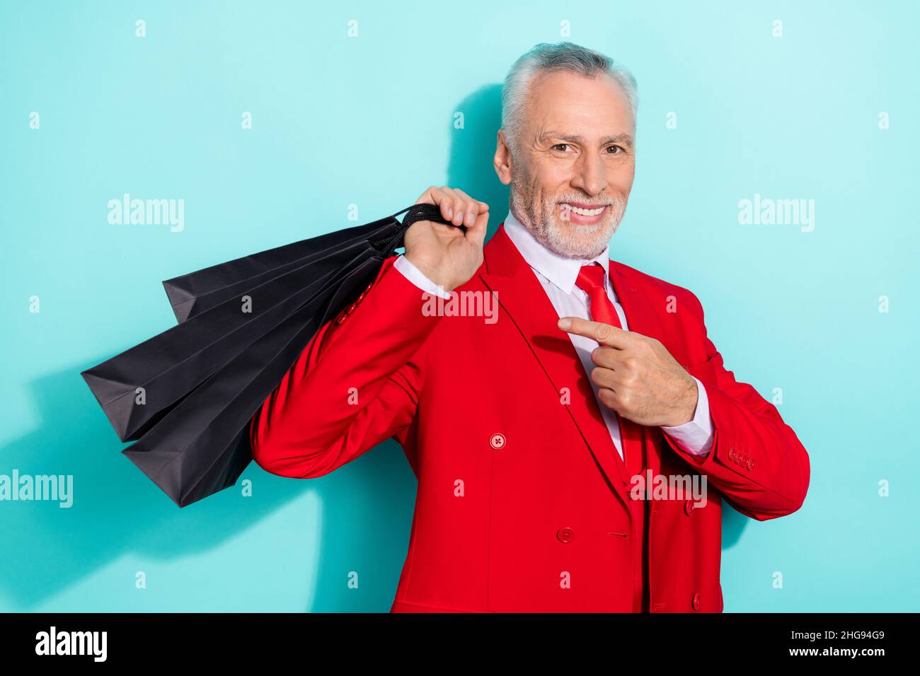 Photo de l'adorable homme retraité porter un costume rouge trois pièces pointant des sacs shopping isolé couleur turquoise arrière-plan Banque D'Images