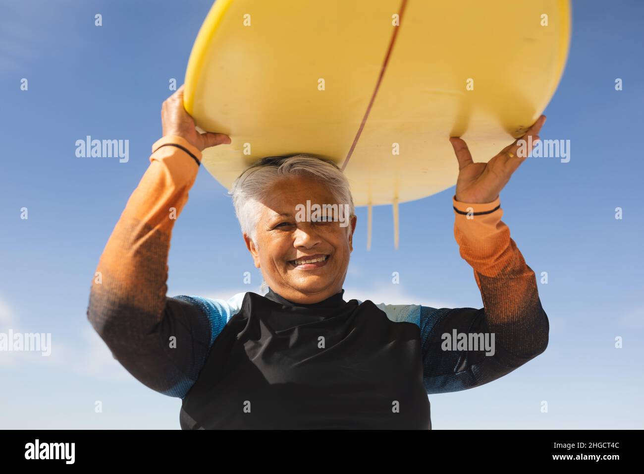 Portrait d'une femme biraciale âgée heureuse portant une planche de surf jaune au-dessus du ciel bleu Banque D'Images