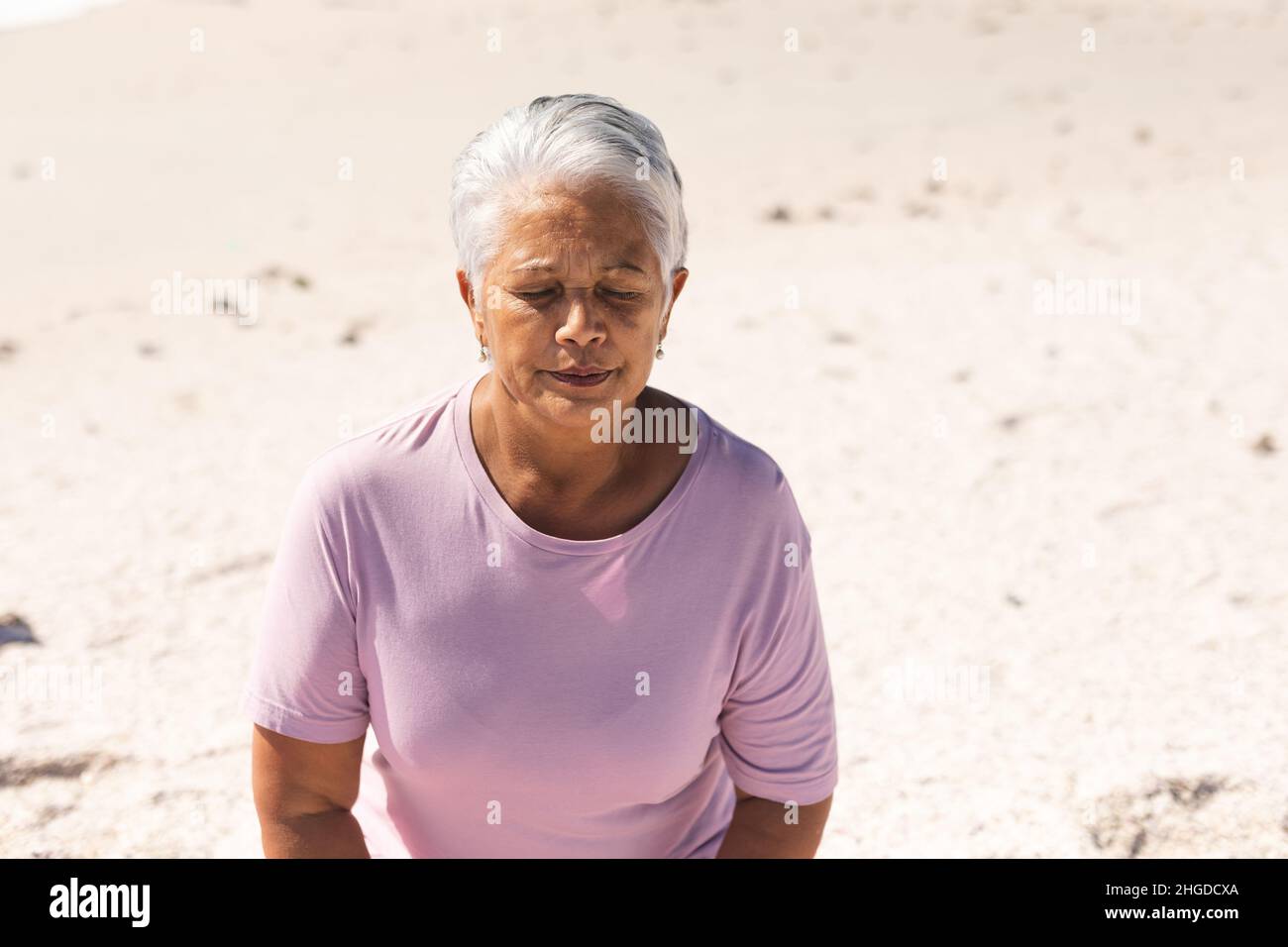 Biracial femme âgée avec de courts cheveux blancs et les yeux fermés méditant à la plage le jour ensoleillé Banque D'Images