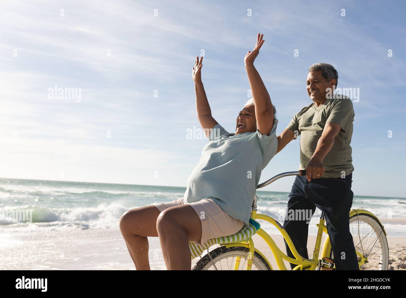 Gaie femme aîée biraciale applaudissante tandis que l'homme à vélo à la plage le jour ensoleillé Banque D'Images