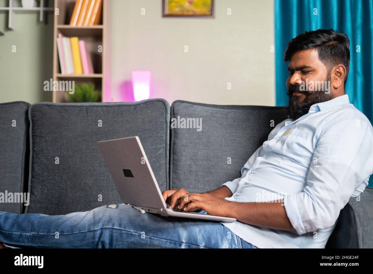 jeune homme souriant, occupé à utiliser un ordinateur portable tout en étant assis sur safa à la maison - concept d'utiliser les médias sociaux, surfer sur le web, travailler à la maison et la technologie Banque D'Images