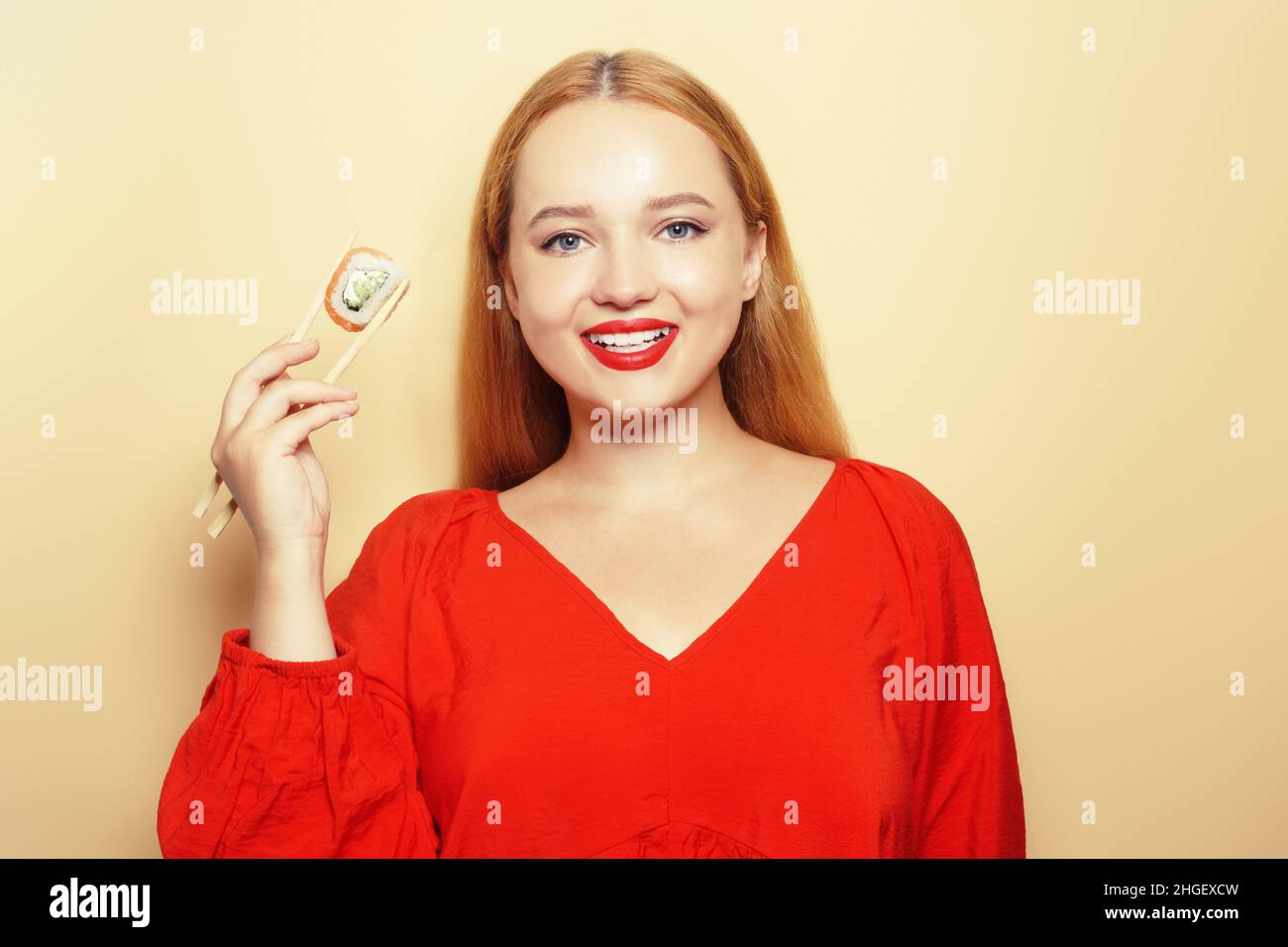 Belle fille mangeant des sushis sur une assiette blanche.Une femme vêtue d'une robe rouge et de lèvres rouges mange des bâtons de sushi avec du saumon de Philadelphie.Modèle souriant et toilettes Banque D'Images