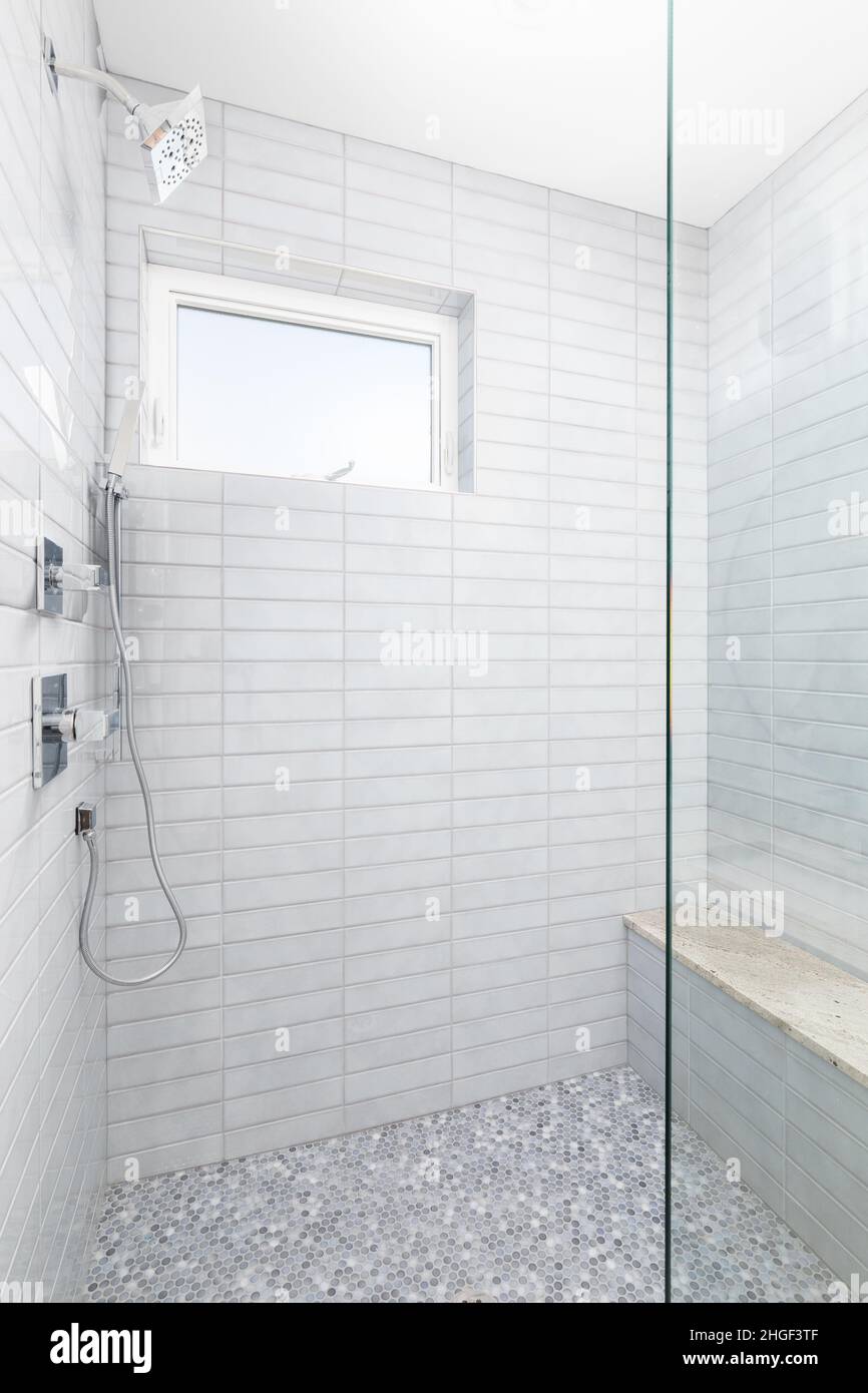 Une douche carrelée grise avec pomme de douche chromée et tuyau, une banquette et de petits carreaux circulaires sur le sol. Banque D'Images