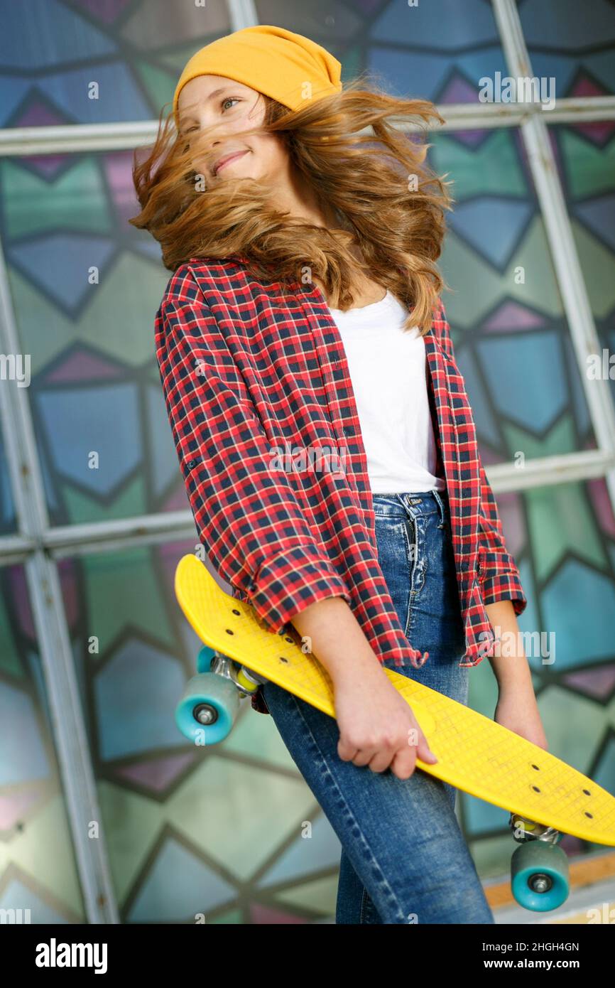 Portrait vertical de style de vie en plein air d'une jeune fille adolescente souriante avec un skateboard jaune portant un chapeau jaune et une chemise à carreaux rouge Banque D'Images