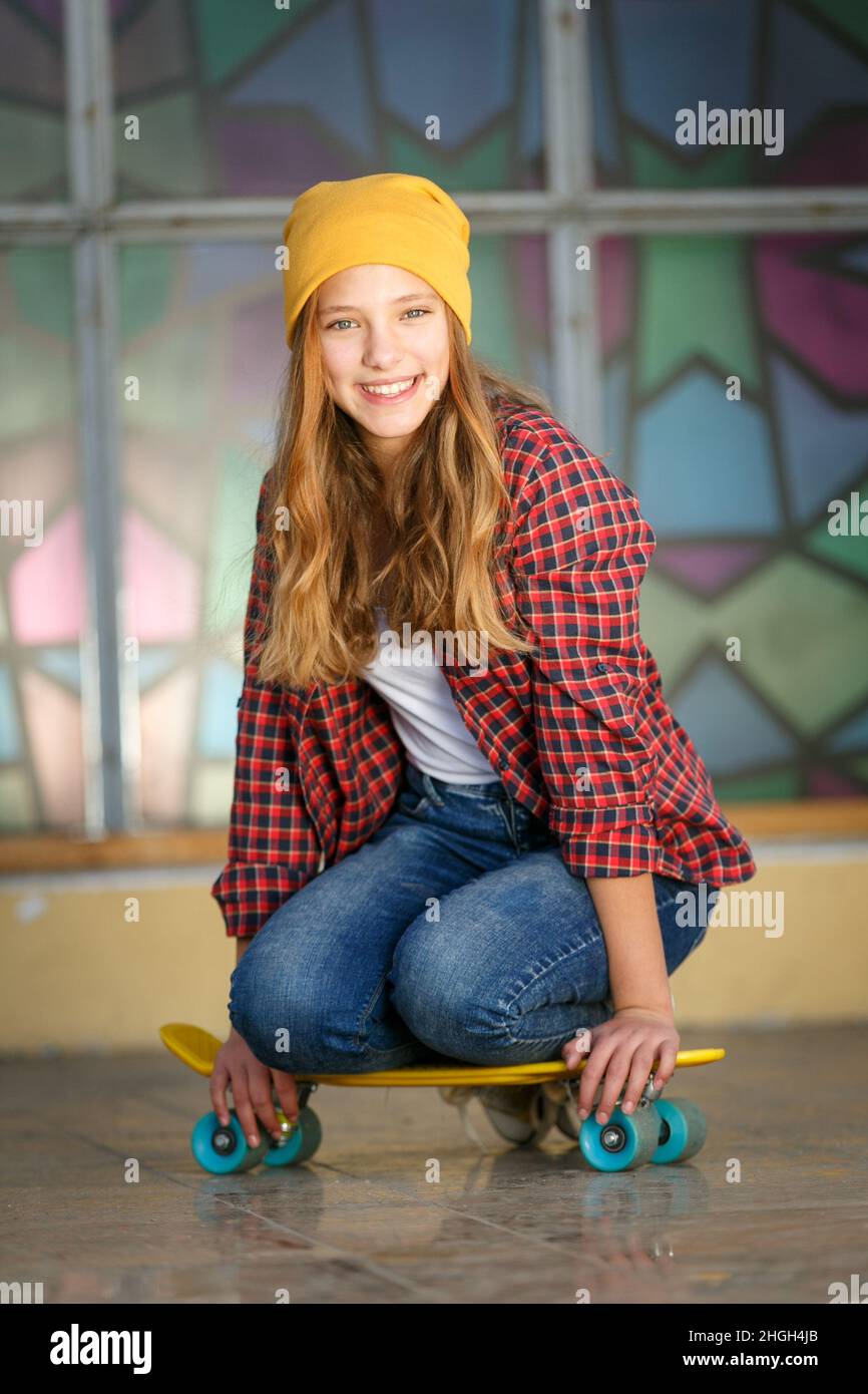 Portrait vertical de style de vie en plein air d'une jeune fille adolescente souriante avec un skateboard jaune portant un chapeau jaune et une chemise à carreaux rouge Banque D'Images