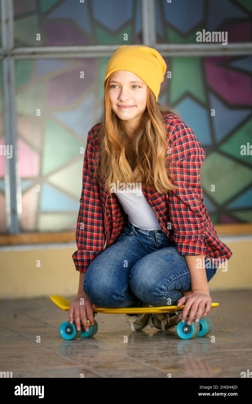 Portrait vertical de style de vie en plein air d'une jeune fille adolescente souriante avec un skateboard jaune portant un chapeau jaune et une chemise à carreaux rouge Banque D'Images
