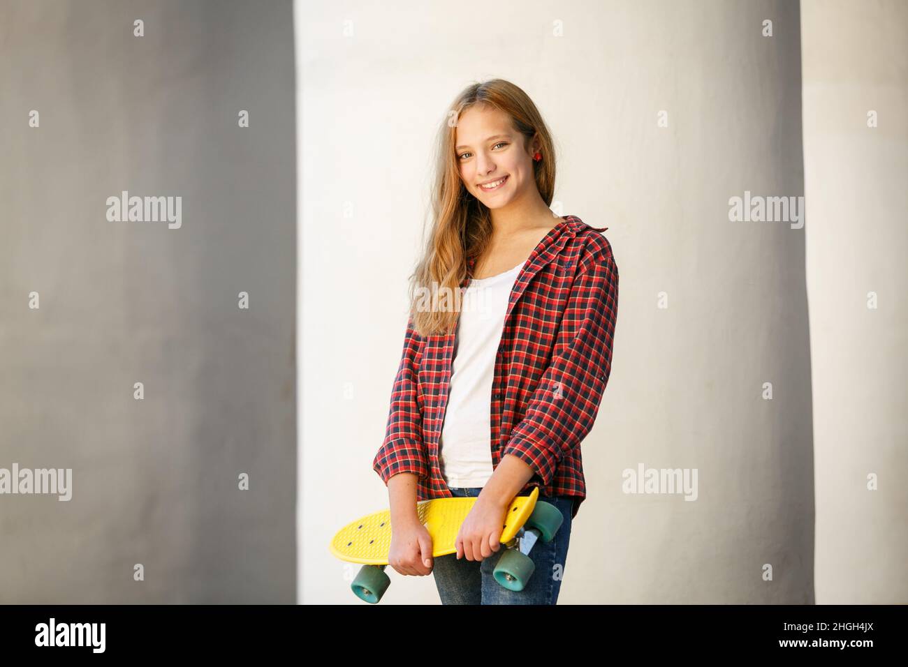 Portrait extérieur horizontal de style de vie d'une jeune fille adolescente souriante avec un skateboard jaune portant une chemise à carreaux rouge Banque D'Images
