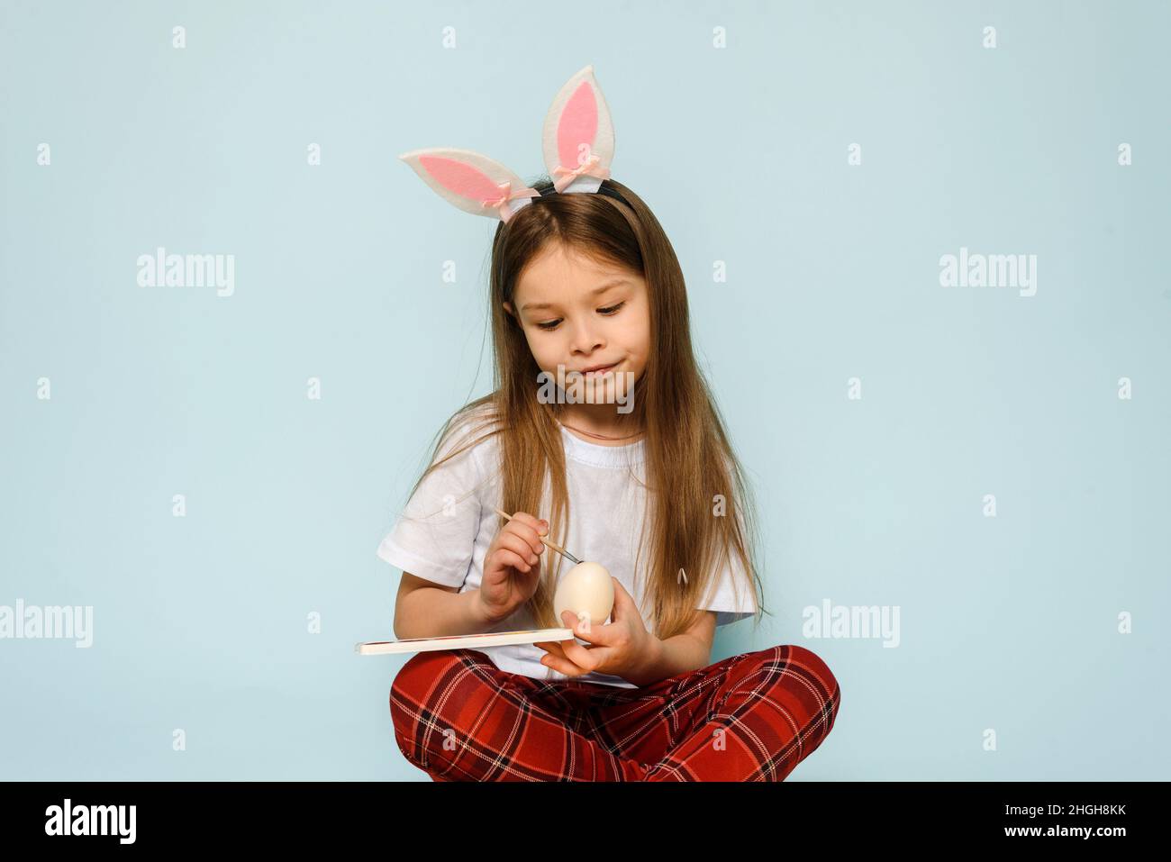 Une petite fille dans les oreilles de lapin peint des oeufs pour Pâques sur un fond bleu dans le studio Banque D'Images