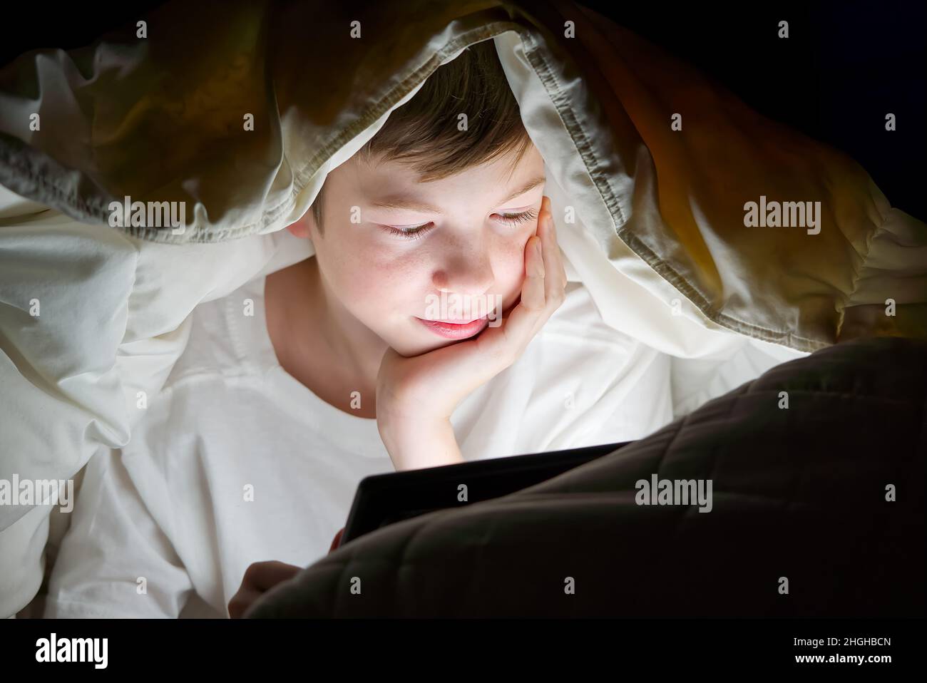 Formation à distance.Formation en ligne.Joyeux garçon dans un lit sous une couverture avec une tablette dans l'obscurité.Le visage de l'enfant est éclairé par un moniteur lumineux.Quarantaine Banque D'Images