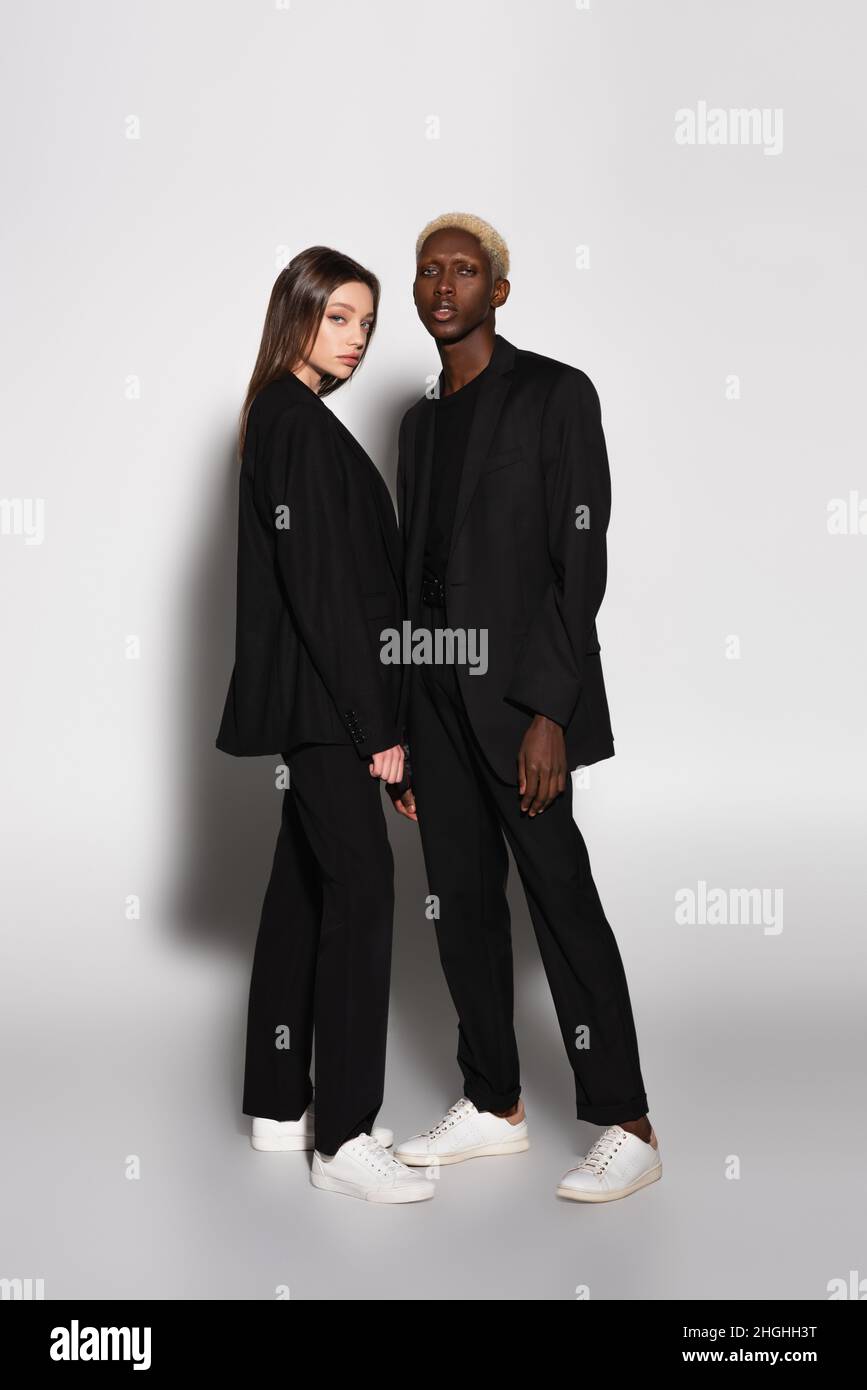 vue sur toute la longueur d'un couple interracial tendance en costume noir et baskets blanches sur fond gris avec ombre Banque D'Images