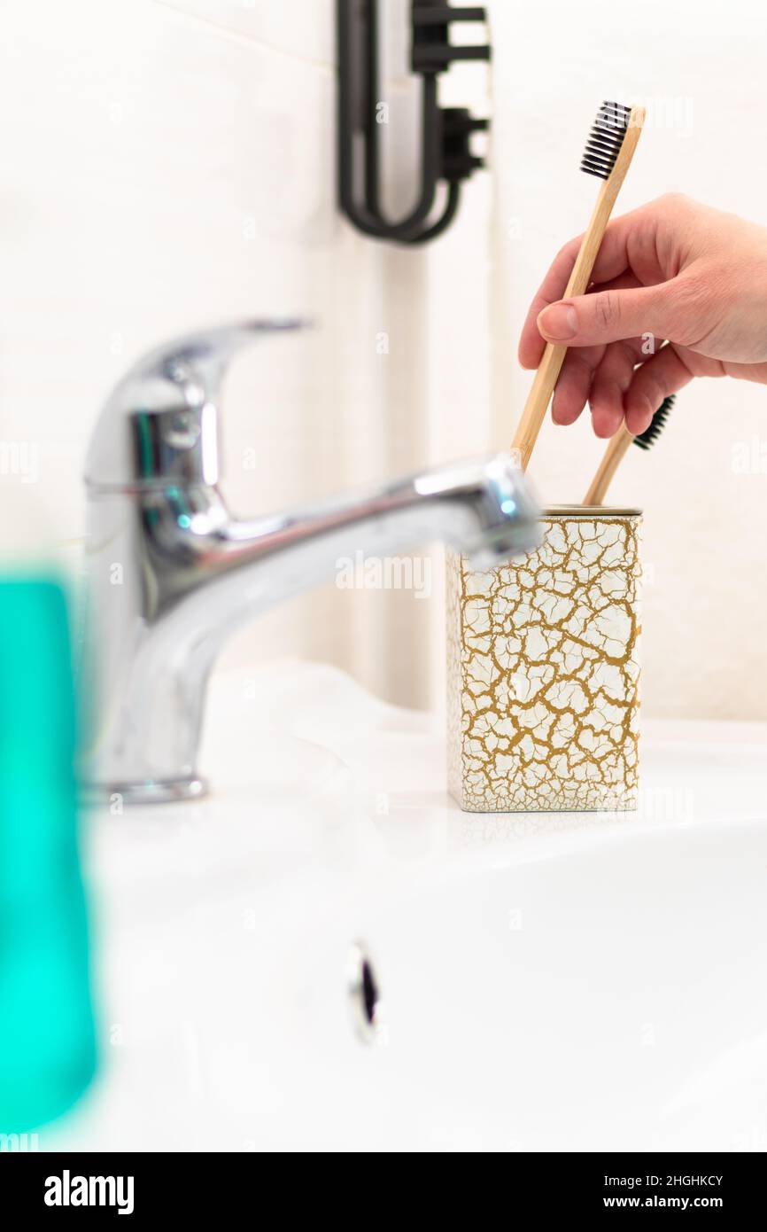 La main d'une femme place une brosse à dents en bois dans un verre avec un motif or dans la salle de bains sur fond d'un robinet et de carreaux brillants.Sélectif Banque D'Images