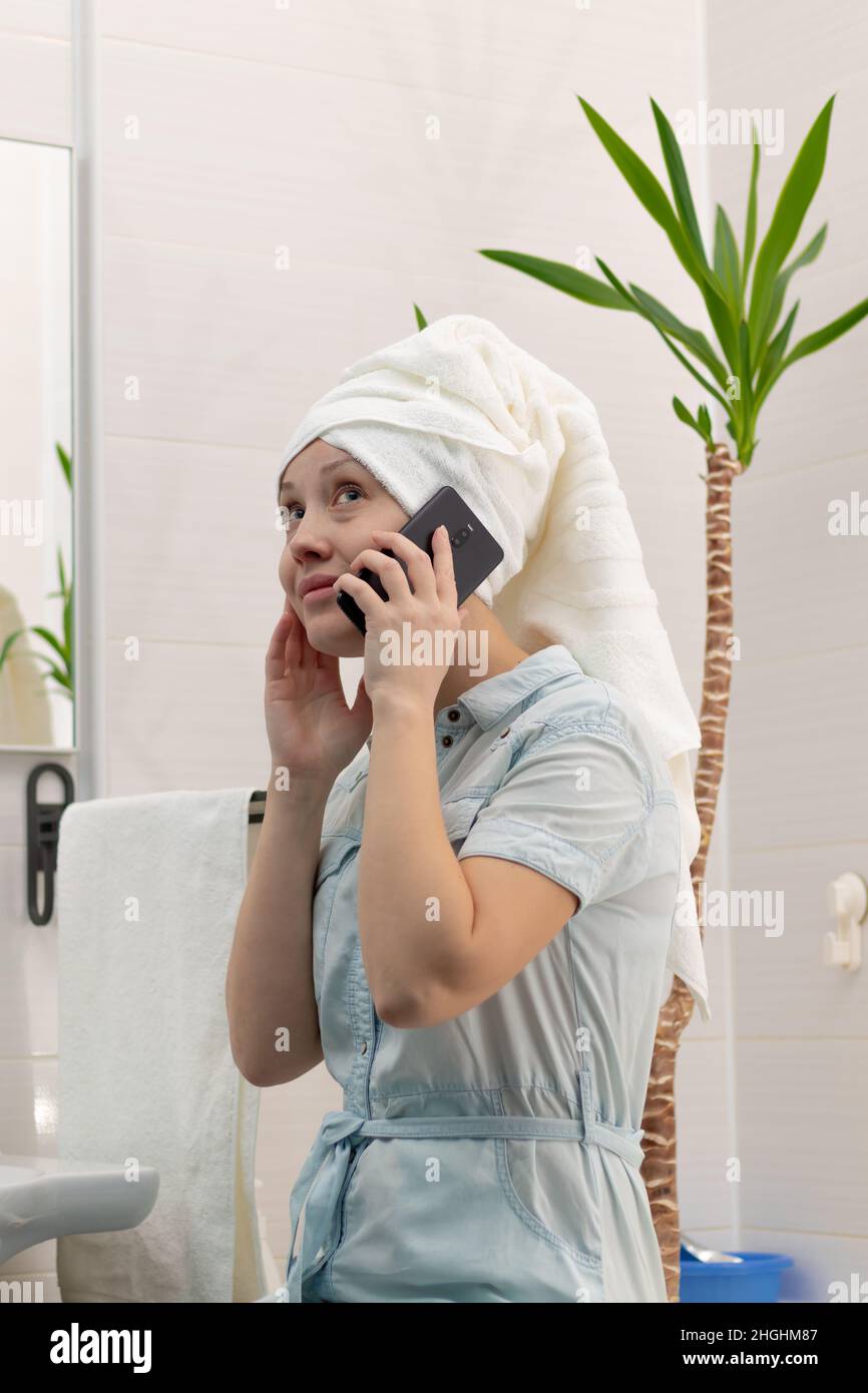 Une jeune femme avec une serviette blanche sur sa tête dans une salle de bains lumineuse communique avec un smartphone avec un ami dans les réseaux sociaux.Réflexion dans le m Banque D'Images