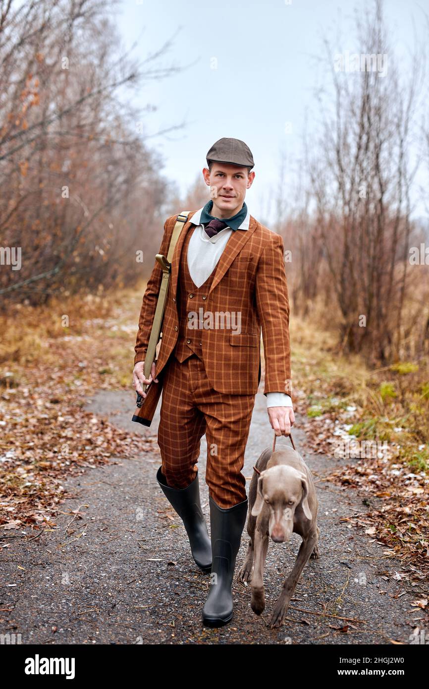 Jeune homme beau chasseur de costume en forêt, homme tenant une arme à feu et marchant avec un chien.Chasseur avec fusil de chasse. Chien puissant et beau en attente Banque D'Images