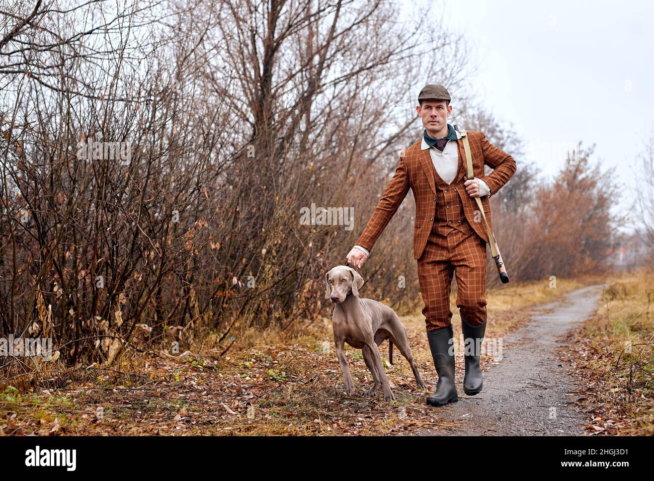 Homme confiant chasseur avec chien de chasse Weimaraner ami portrait en zone rurale pendant la saison de chasse, beau type caucasien en costume brun, à pied Banque D'Images