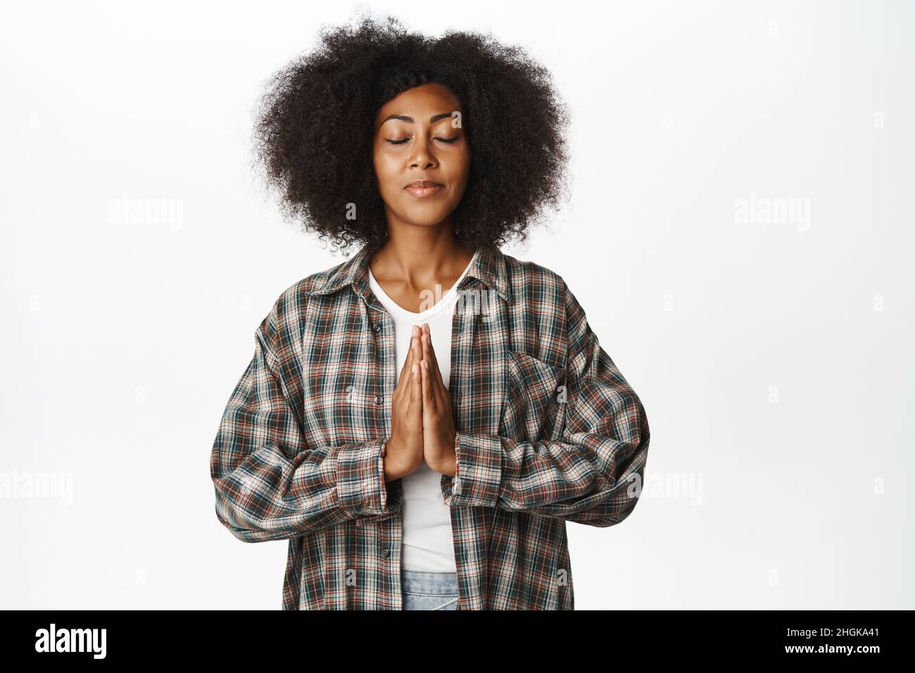 Femme africaine pacifique et consciente priant, tenant les mains dans la prière, geste namaste et les yeux proches, se détendant et respirant librement, debout sur blanc Banque D'Images