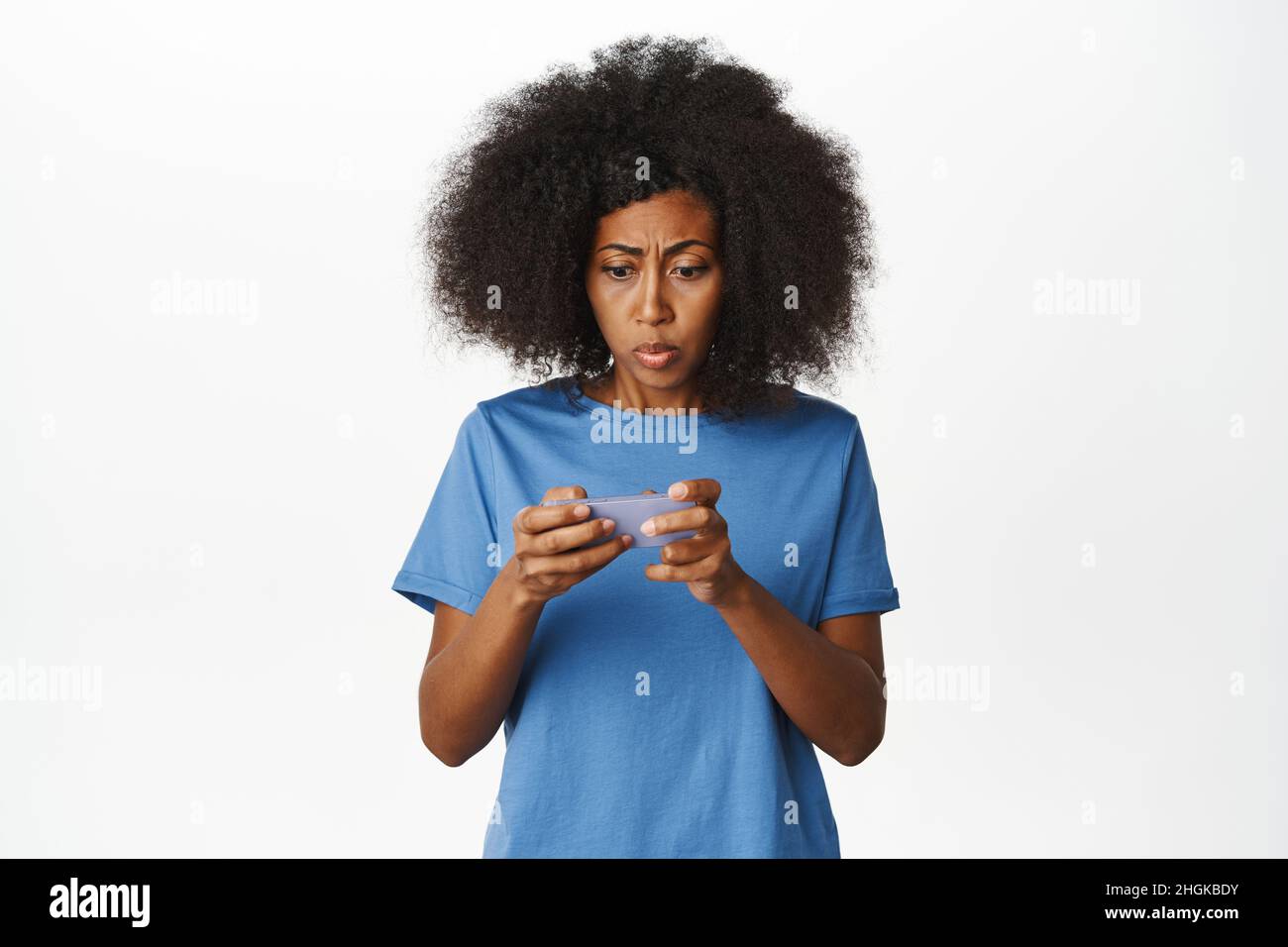 Femme regarde sérieusement l'écran horizontal de téléphone mobile, femme afro-américaine regardant la vidéo sur l'application de smartphone avec l'expression concernée, blanc Banque D'Images
