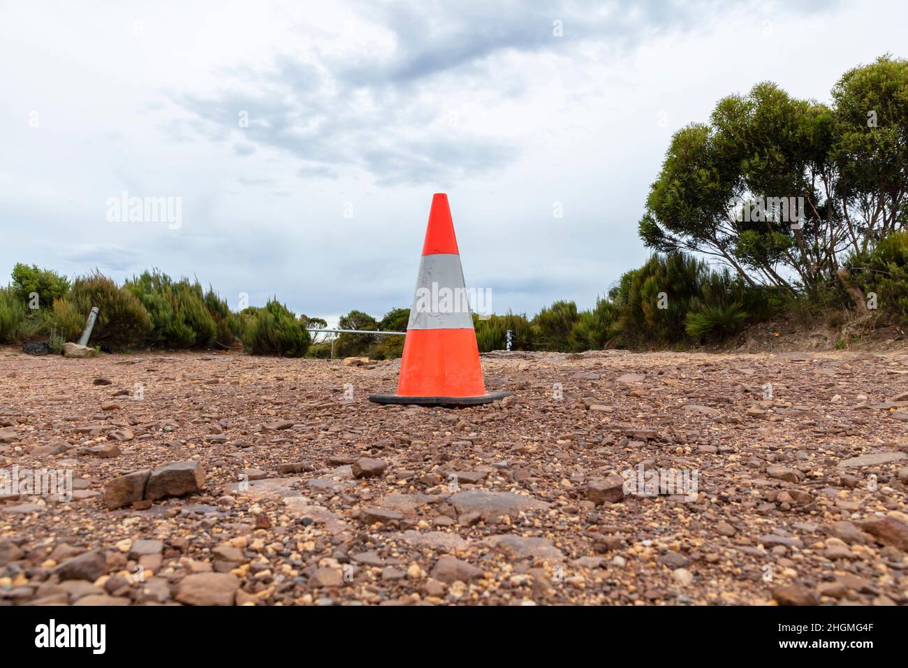 Photographie d'un cône de sécurité orange ancien et endommagé avec du ruban fluorescent déchiré sur une route rocheuse en terre devant les arbres et une clôture de câble Banque D'Images