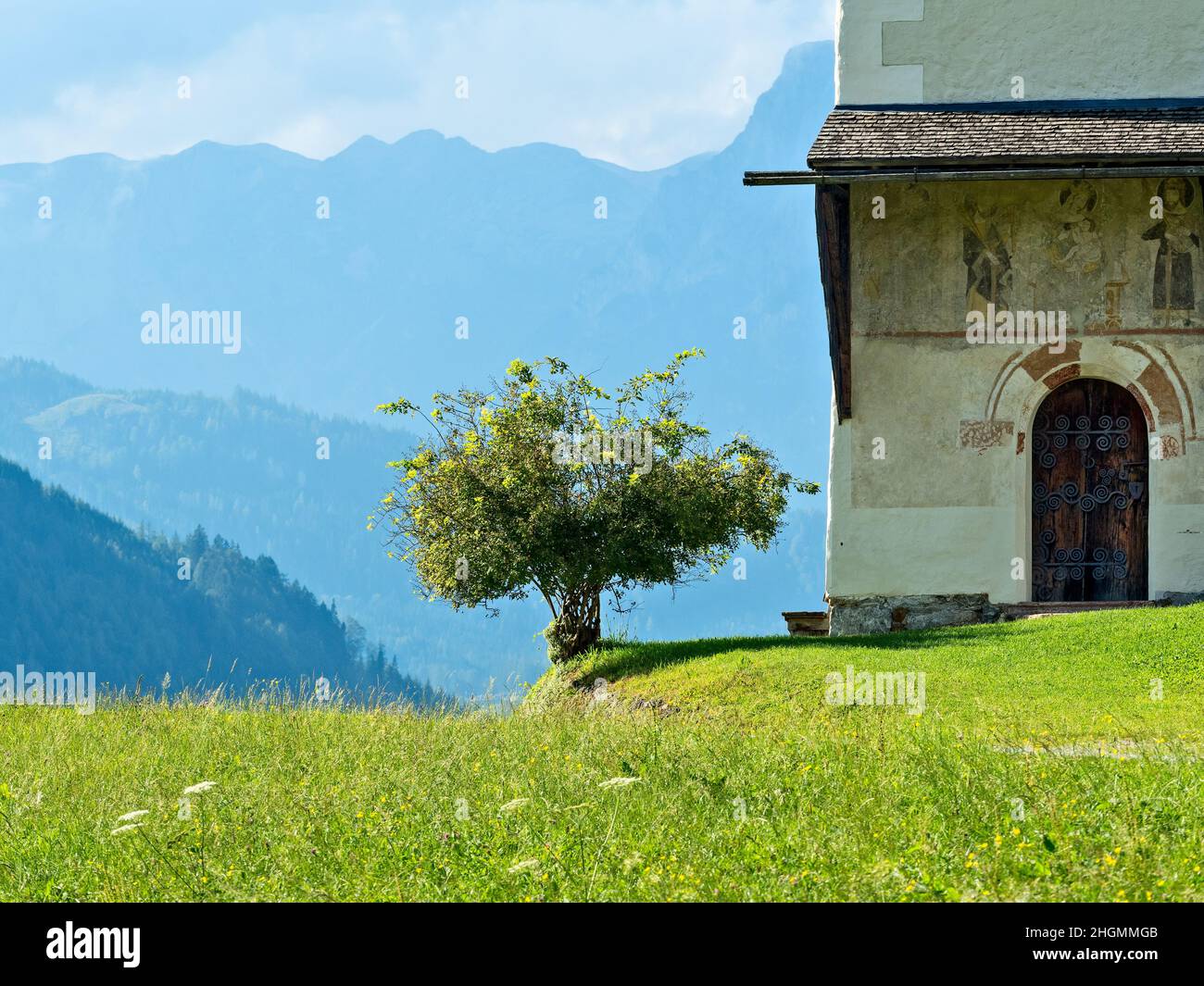 Un pré d'été avec un ciel bleu et quelques nuages blancs avec une grande chaîne de montagnes en arrière-plan.Une petite chapelle avec un arbre fruitier peut être Banque D'Images