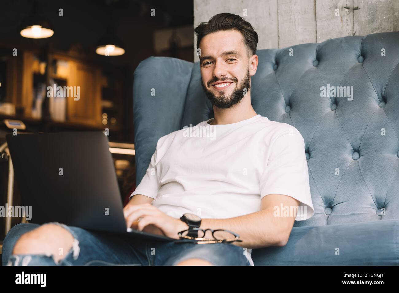 Portrait d'un homme hipster qualifié souriant à l'appareil photo tout en faisant un travail à distance d'ordinateur portable dans l'espace de travail de collègue Banque D'Images