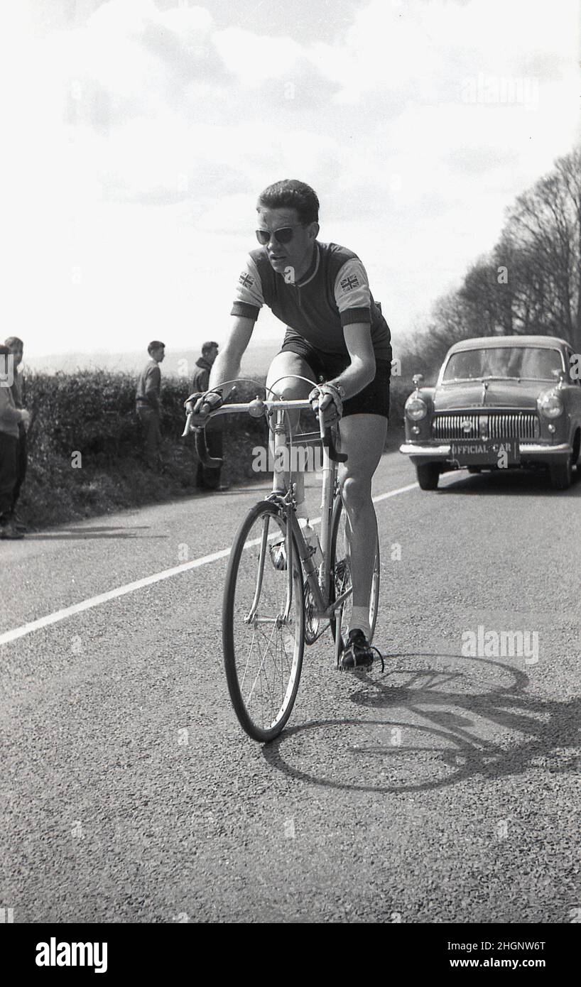 1950s, bisisotrique, cycliste professionnel en compétition dans une course sur route en face de la voiture de course officielle, Angleterre, Royaume-Uni.Les badges Union Jack sur les épaules de son maillot de course, mais pas le nom ou le logo du sponsor. Banque D'Images