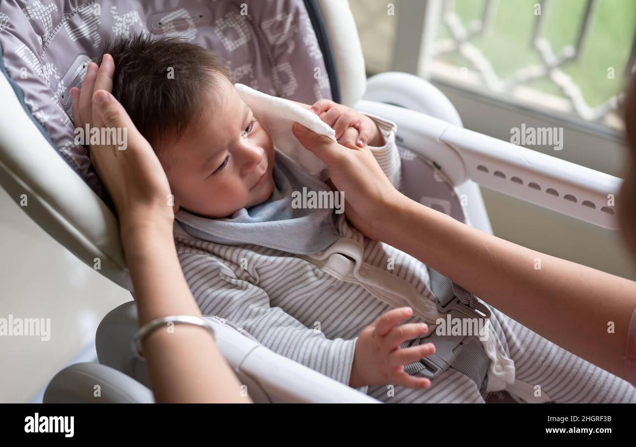Mère essuyant le visage de son bébé garçon avec un papier de soie de coton alors qu'il est assis sur une chaise haute après avoir pris un repas près de la fenêtre à la maison Banque D'Images