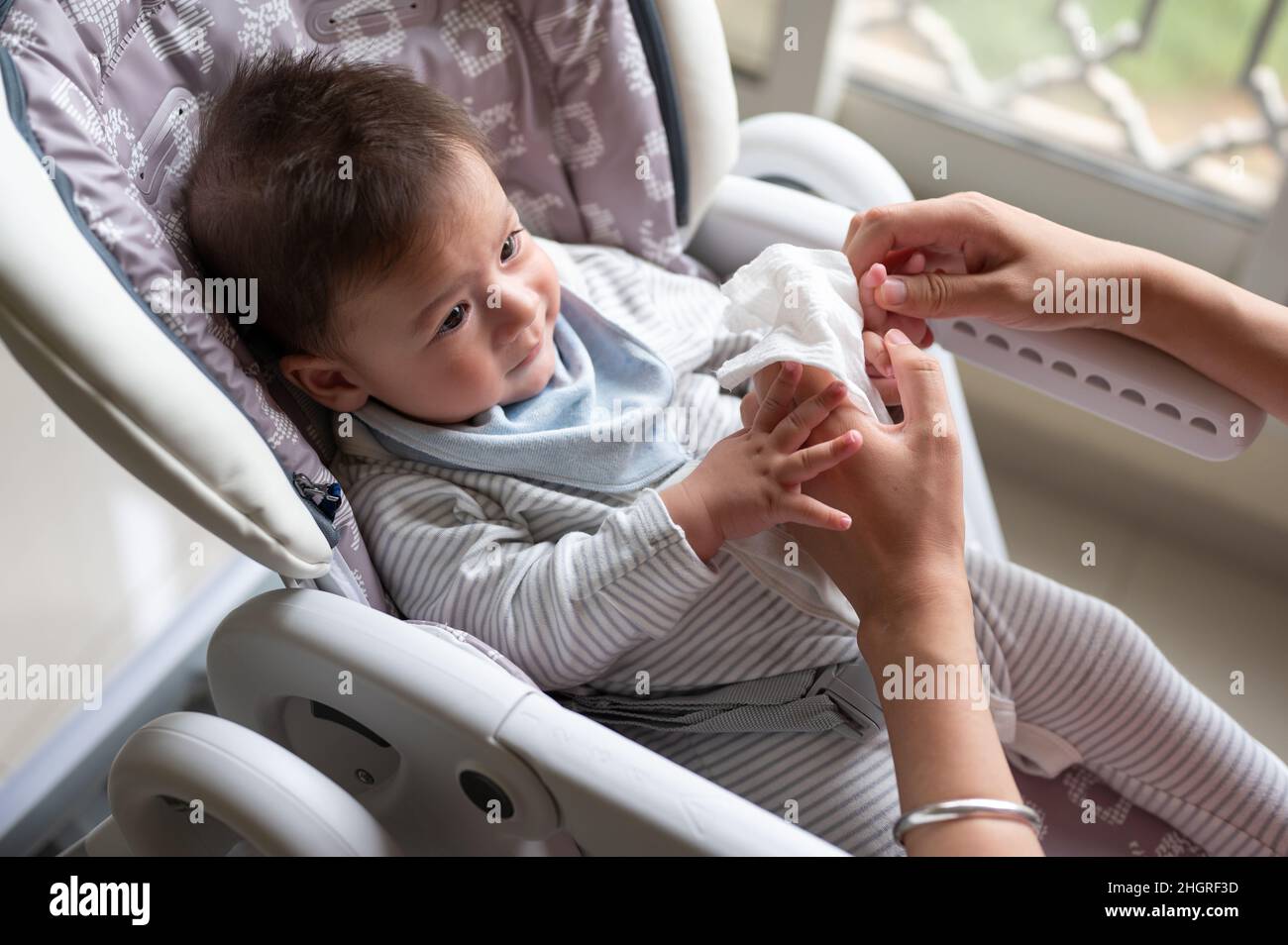 Mère essuyant le visage de son bébé garçon avec un papier de soie de coton alors qu'il est assis sur une chaise haute après avoir pris un repas près de la fenêtre à la maison Banque D'Images