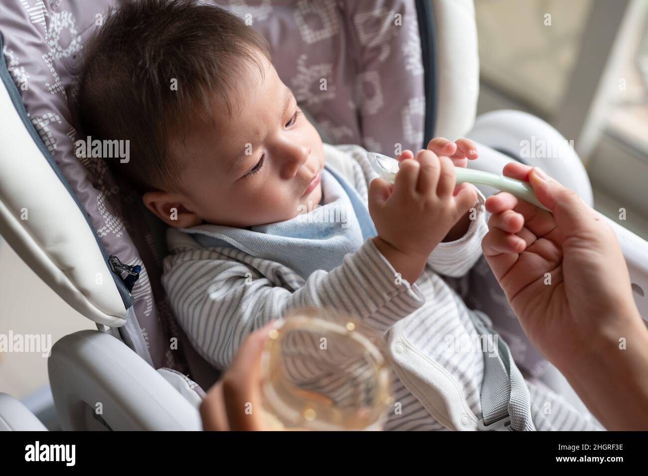 Mère nourrissant l'eau de son bébé garçon avec une cuillère dans une chaise haute près de la fenêtre à la maison Banque D'Images