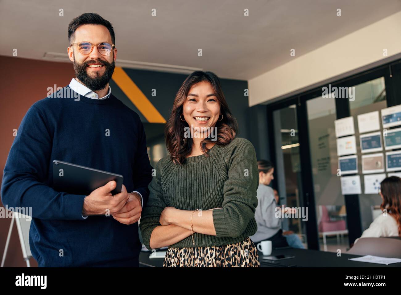 Deux entrepreneurs heureux souriant à la caméra joyeusement.Jeunes hommes d'affaires debout dans une salle de réunion avec leurs collègues en arrière-plan.Plongeurs Banque D'Images