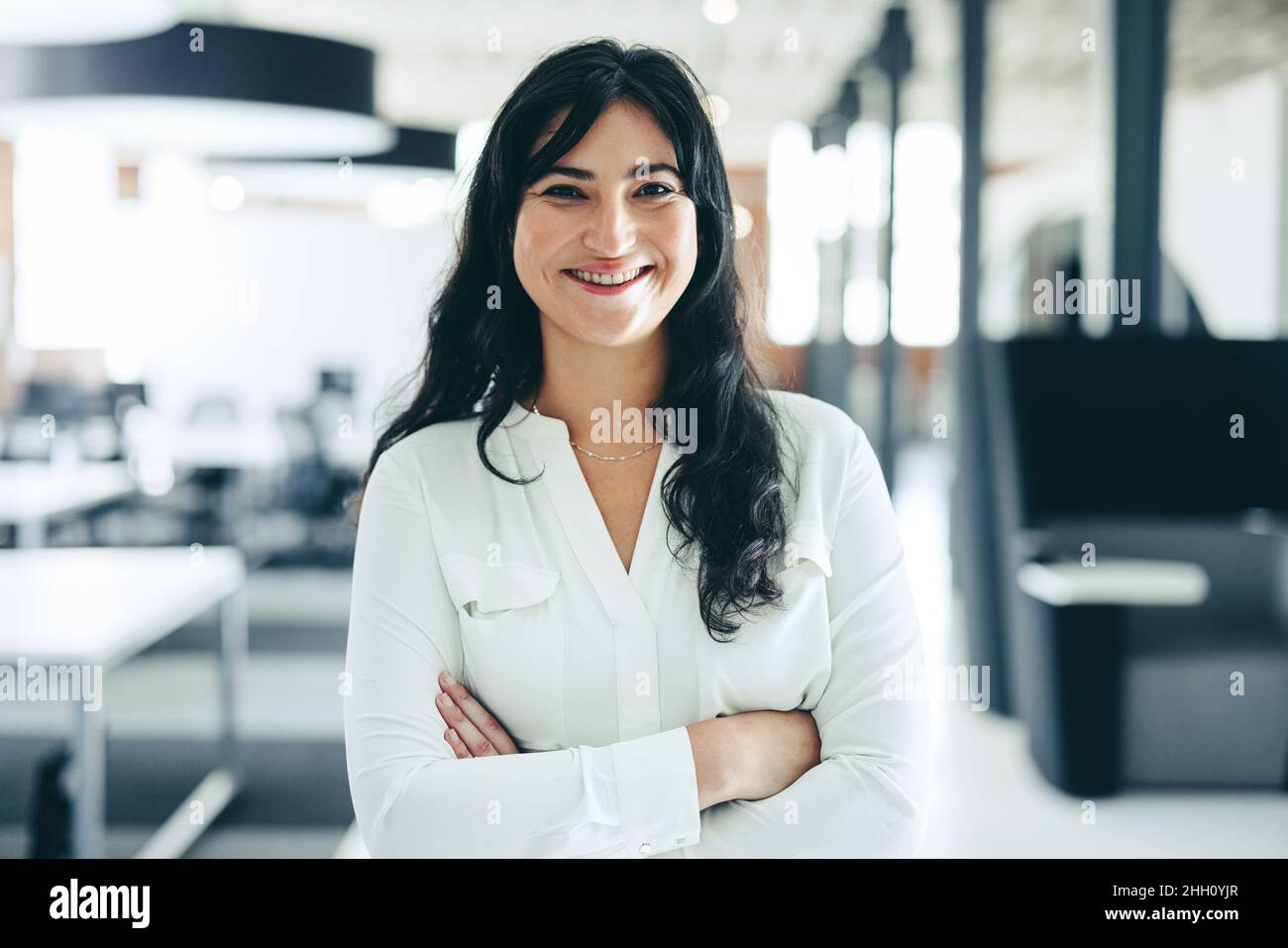Femme d'affaires souriante à la caméra.Une jeune femme d'affaires heureuse debout avec ses bras croisés dans un milieu de travail moderne.Femme d'affaires joyeuse Banque D'Images