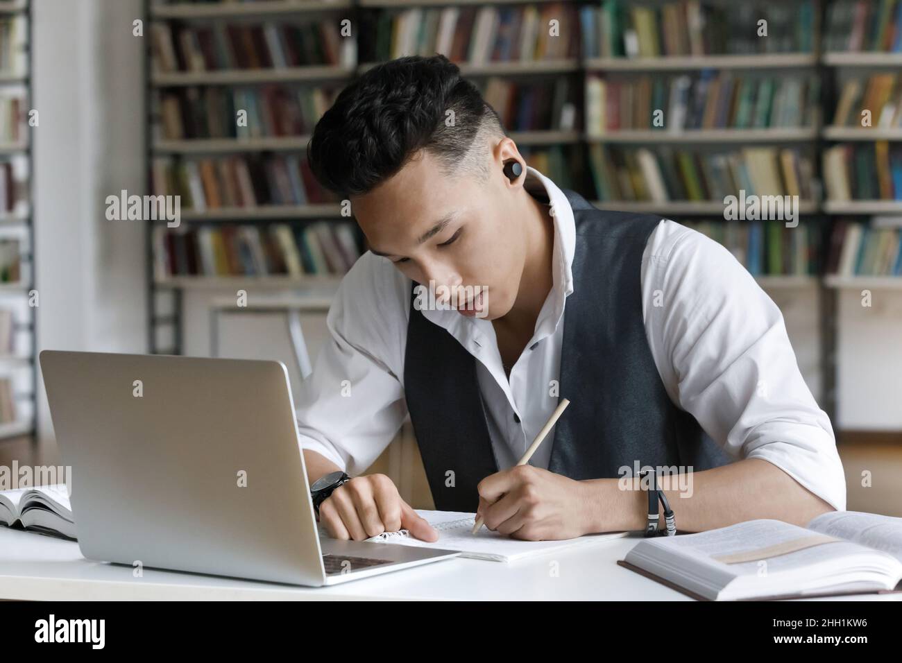 Un étudiant asiatique sérieux et concentré avec des écouteurs sans fil pour écrire des notes Banque D'Images