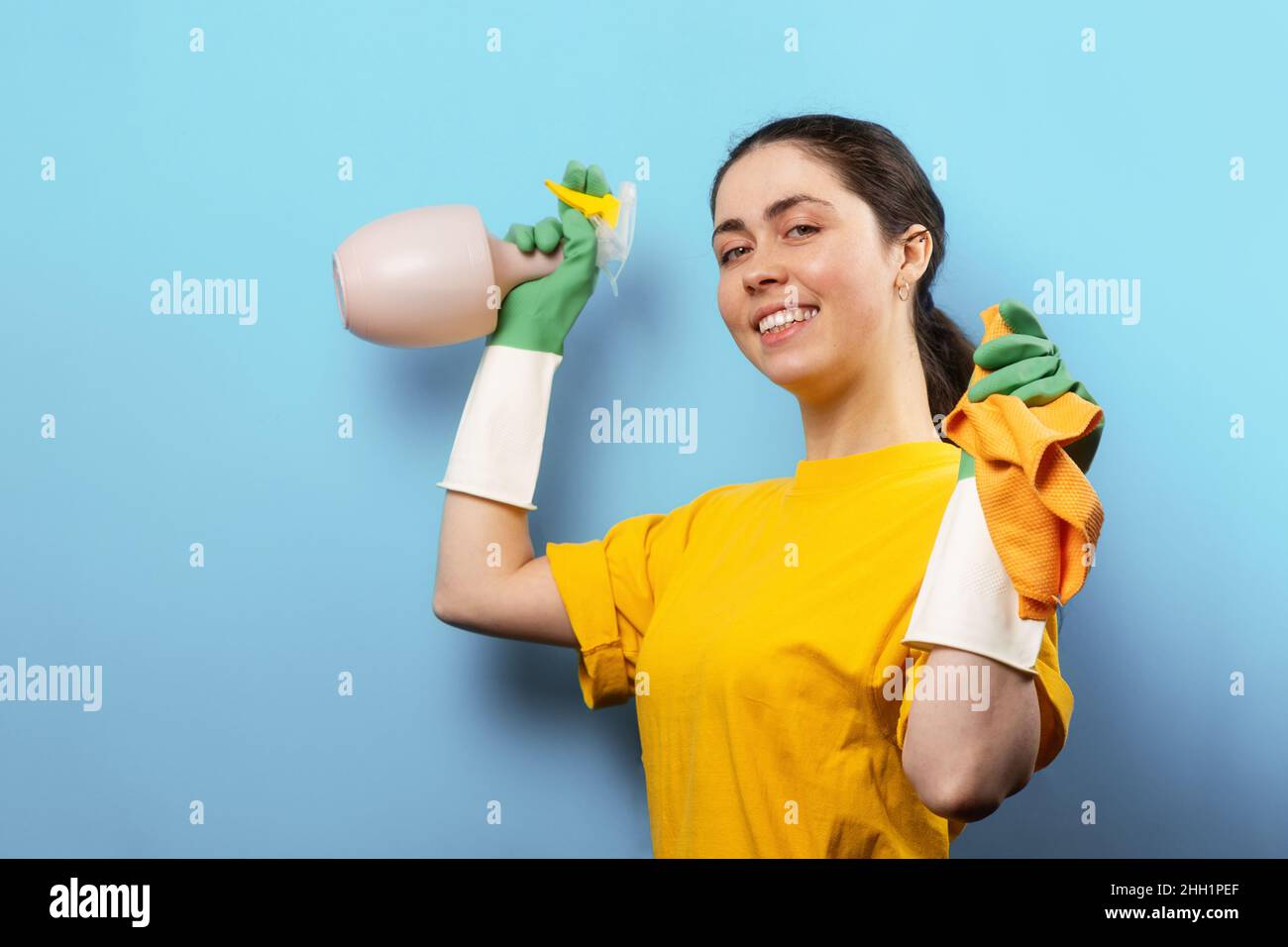 Portrait d'une jeune femme caucasienne souriante en gants de caoutchouc, tenant un pulvérisateur et un chiffon dans les mains.Arrière-plan bleu avec espace de copie.Concept de clea Banque D'Images
