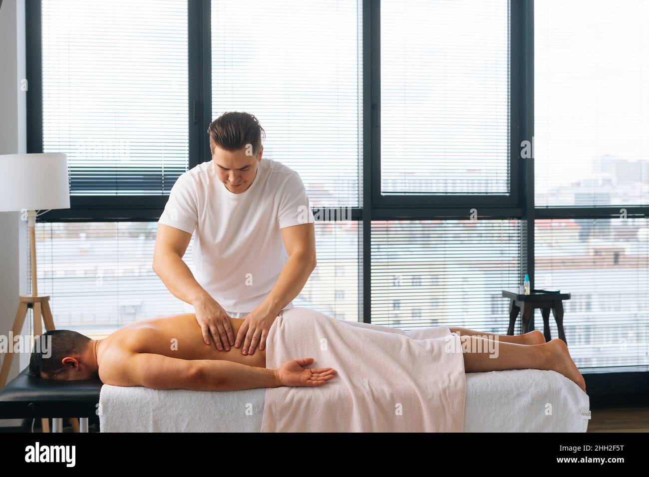 Prise de vue large d'un masseur masculin professionnel avec des mains fortes, massant le côté du dos à un sportif musclé allongé sur l'estomac, Banque D'Images