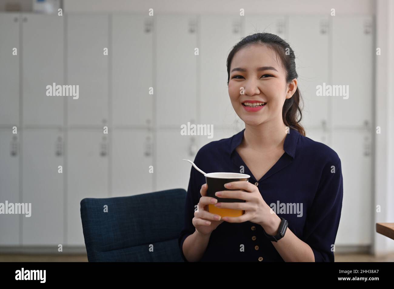 Femme asiatique joyeuse tenant une tasse de nouilles instantanées et souriant à l'appareil photo. Banque D'Images