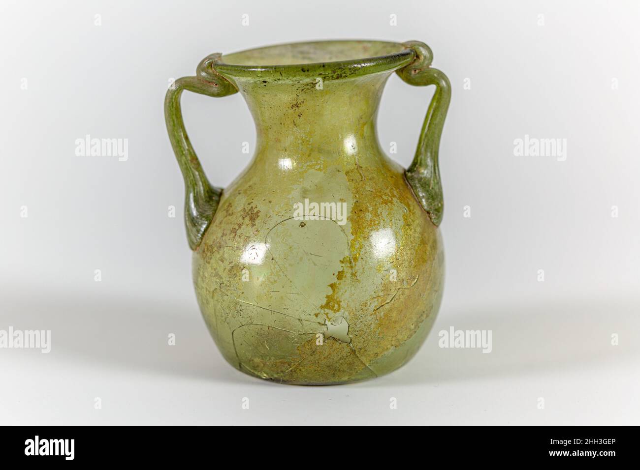 Pot en verre à deux poignées vert translucide romain 4th Century A.D. ; poignées de la même couleur. Rebord arrondi légèrement épaissi ; col évasé large ; col concave ; corps bulbeux ;fond poussé avec cicatrice de pontil centrale ;Deux poignées de tige appliquées de manière inégale sur le dessus du corps dans de grands coussins, orientés vers le haut et vers l'extérieur, puis tournées vers l'intérieur et pliées sur le dessous de la bouche et sur le bord, se terminant sur la lèvre intérieure. Cassées et réparées d'un côté, avec un petit trou d'impact et de grandes fissures;quelques bulles plus grosses et plus petites, avec des impuretés noires autour de la bouche et dans les poignées; légère morcellement et légère iridescence à l'extérieur, taches Banque D'Images
