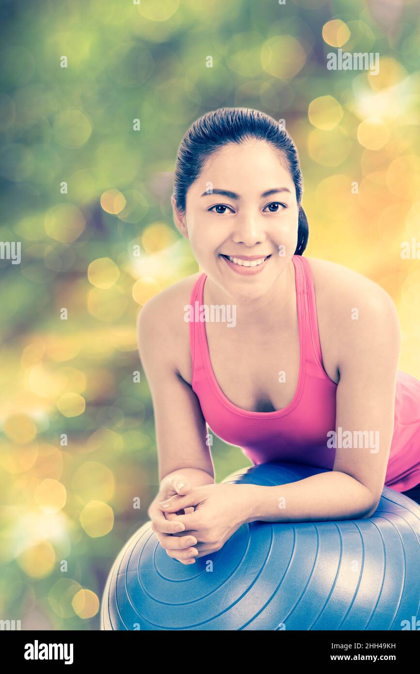 Femme en bonne santé - belle fille asiatique (thaï) souriant et penchée sur une balle de fitness dans le studio de fitness, scintillant lumières flou bokeh lumière abstraite spr Banque D'Images