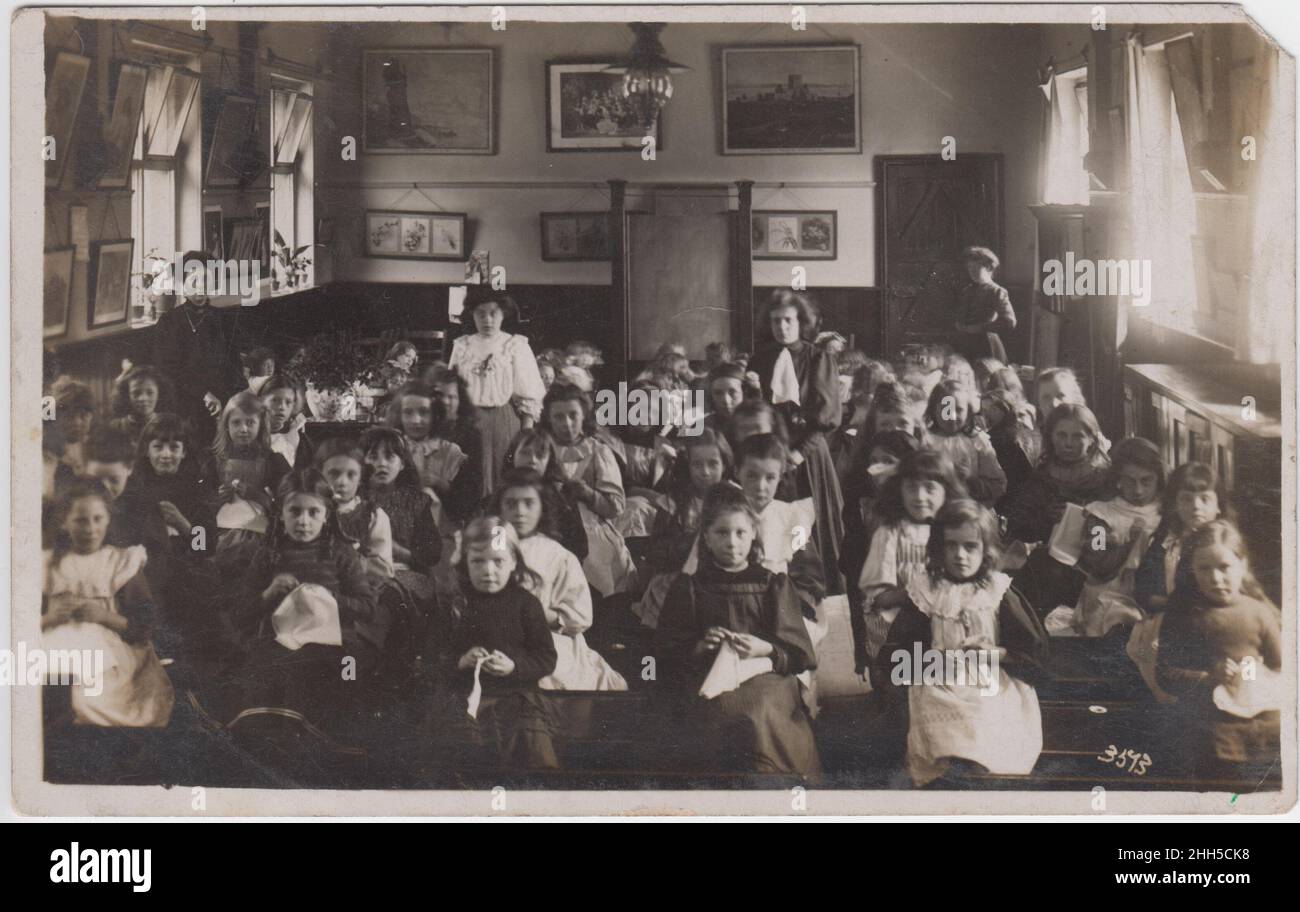 Les filles assis dans une salle de classe, apprenant la broderie.Quatre enseignants sont debout dans la salle de l'école, qui est décorée avec beaucoup d'images.Photographie prise au début du 20th siècle Banque D'Images