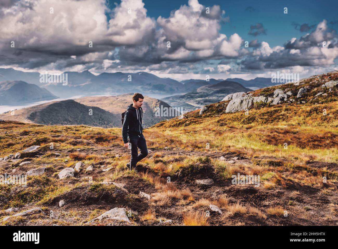 Jeune homme adulte en randonnée au sommet d'une montagne en norvège pendant l'automne, portant un sac à dos, regardant vers le bas, le ciel et l'horizon avec la chaîne de montagnes, volda norw Banque D'Images