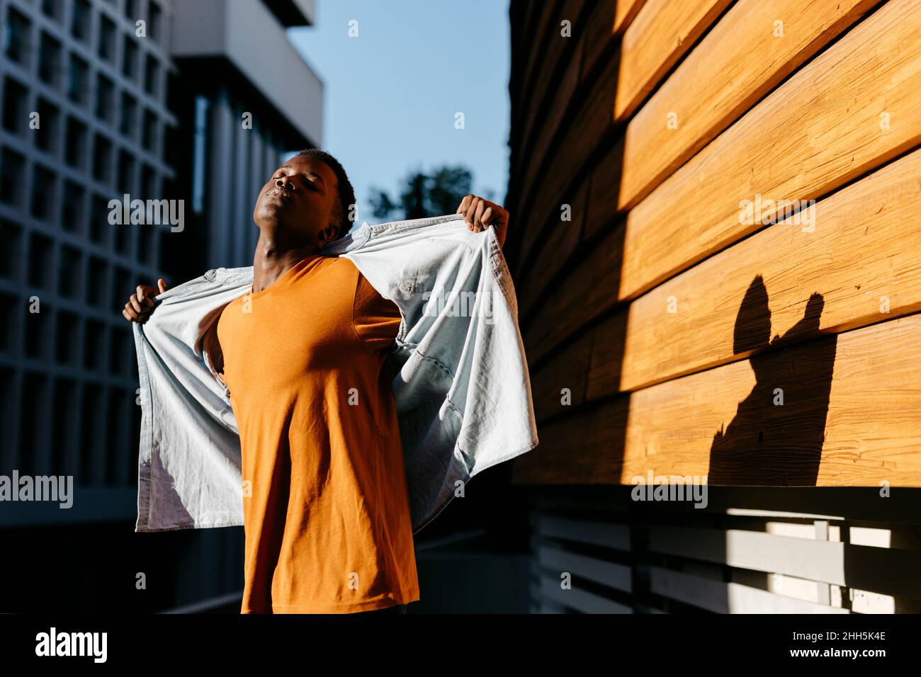 Jeune homme avec les yeux fermés tenant une chemise ouverte par le mur au coucher du soleil Banque D'Images
