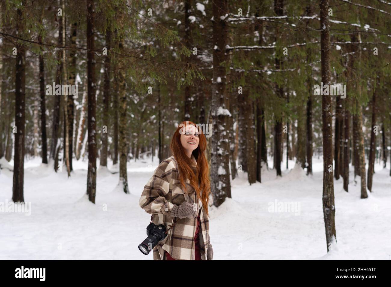 Femme souriante à tête rouge avec caméra jour rêvant dans la forêt d'hiver Banque D'Images