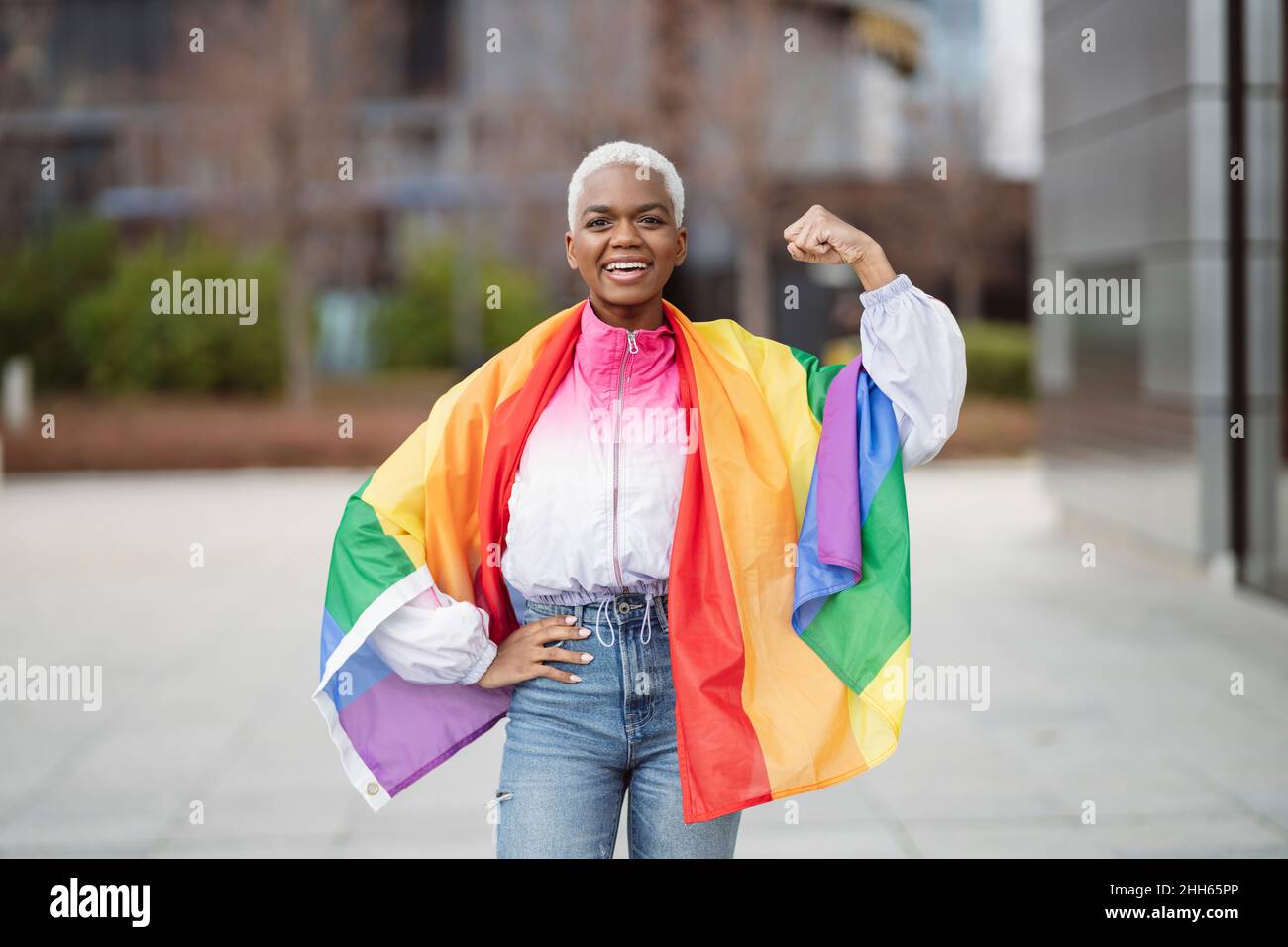 Jeune femme enveloppée dans un drapeau arc-en-ciel fléchissant le muscle avec la main sur la hanche Banque D'Images