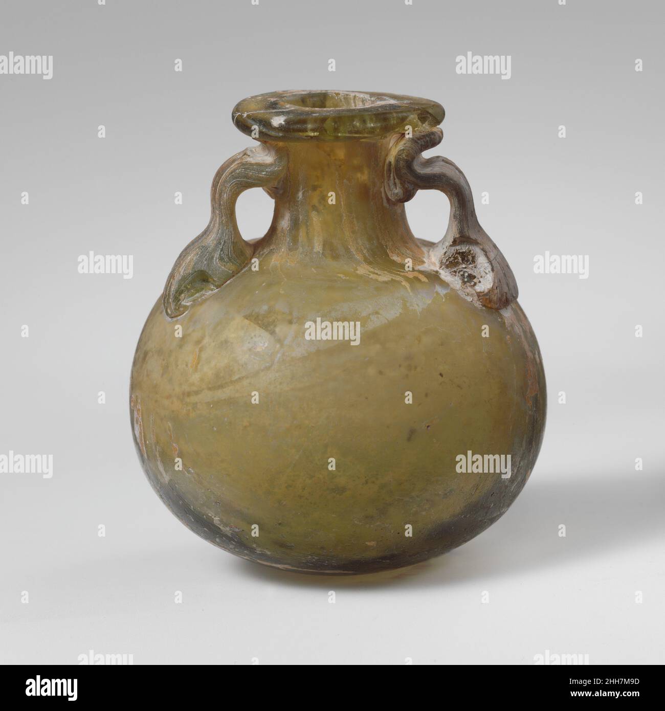 Aryballos en verre (bouteille d'huile) mi-1st–3rd Century A.D. fond d'huile romain à parois épaisses vert translucide jaune-translucide; poignées de la même couleur.rebord replié, sur-dessus et à l'intérieur et aplati sur le dessus; col cylindrique, évasant à la base pour joindre le squat, corps globulaire; fond arrondi;Deux poignées appliquées sur le dessus du corps dans de grands coussins, tracées, pliées et pressées sur le dessus du cou et le dessous de la jante.corps intact, mais une poignée cassée et réparée; une grande puce manquante dans la jante; puce avec des bris manquants dans le coussinet inférieur de chaque poignée;quelques bulles et inclusions vitreux; morcellement, piqûres et irid Banque D'Images