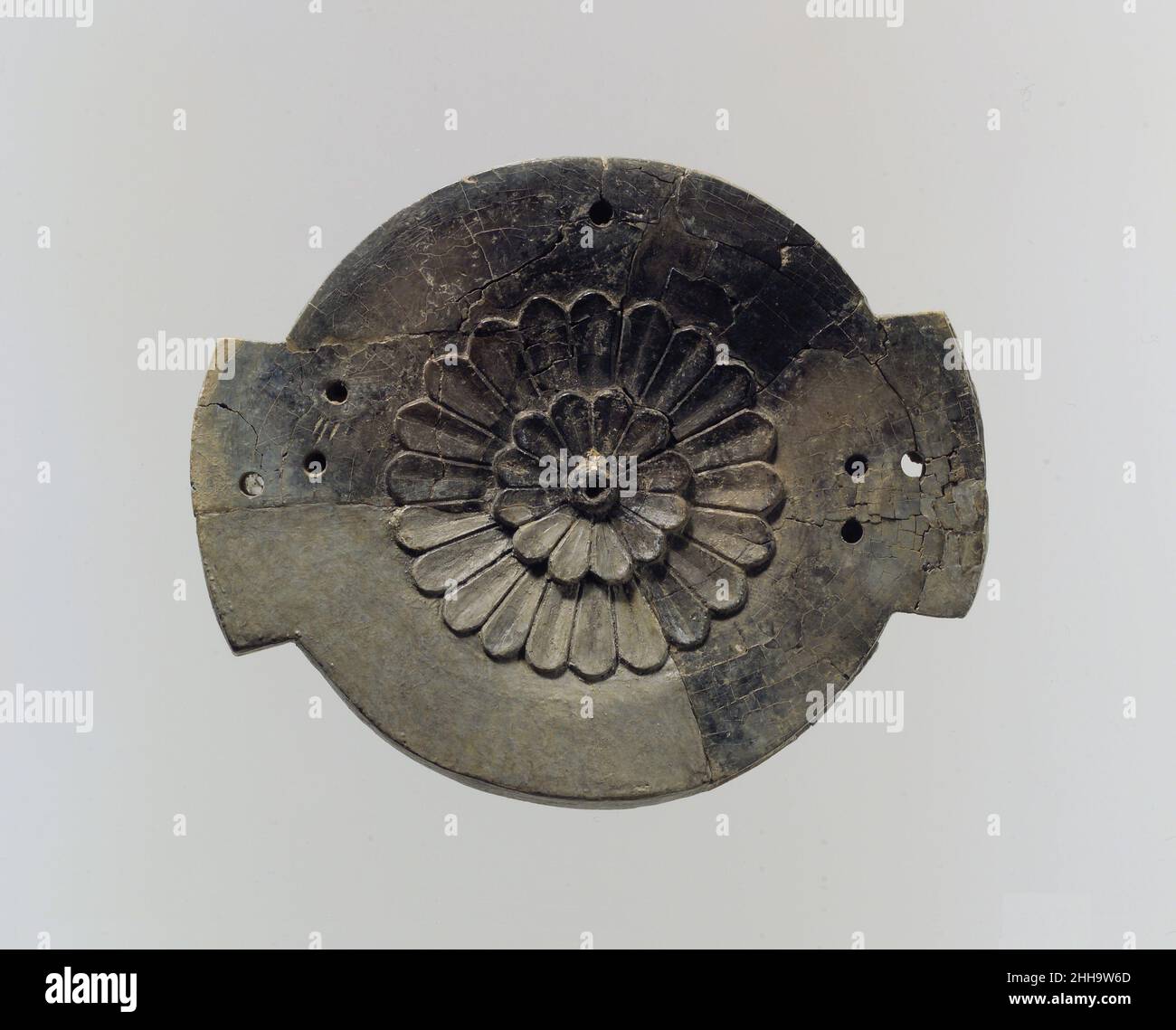 Fragment de couvercle ca.9th–8th siècle av. J.-C. Assyrian ce morceau brisé d'ivoire sculpté était probablement un couvercle pour une petite boîte ronde, appelée pyxis par des chercheurs.Environ un quart du couvercle a été restauré après l'excavation de l'objet et peut être distingué de l'objet ancien par sa couleur uniforme et sa texture de surface.La surface supérieure du couvercle est décorée d'une rosette centrale avec deux rangées concentriques de pétales.Le couvercle a probablement été fixé à sa boîte par des chevilles placées à travers les trous percés à travers les deux languettes symétriques projetant du bord, ce qui permet au couvercle de pivoter ouvert quand un Banque D'Images