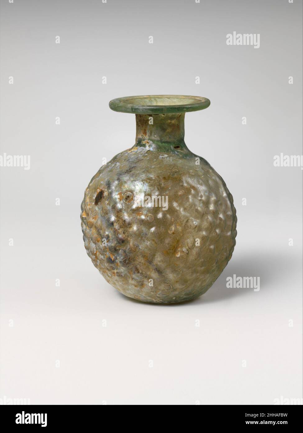 Fiole sprinkleur en verre 4th Century A.D. Roman translucide vert pâle avec teinte bleue. Large rebord horizontal replié, vers le bas, rond et intérieur ; col cylindrique,avec constriction à la base ; corps globulaire ; petit fond concave.Remorquer les coutures proéminentes du moule sur les côtés de la carrosserie. Sur la carrosserie, le motif de losange se chevauchant des petits boutons relevés, indistincts en haut et en bas. Intact, sauf pour deux petits trous dans le corps; quelques bulles;morcellement, piqûres de grosses bulles de surface, iridescence, et taches d'intempéries incrustées à l'extérieur, enorquage et brillant irisé à l'intérieur.Arroseur en verre, fl Banque D'Images