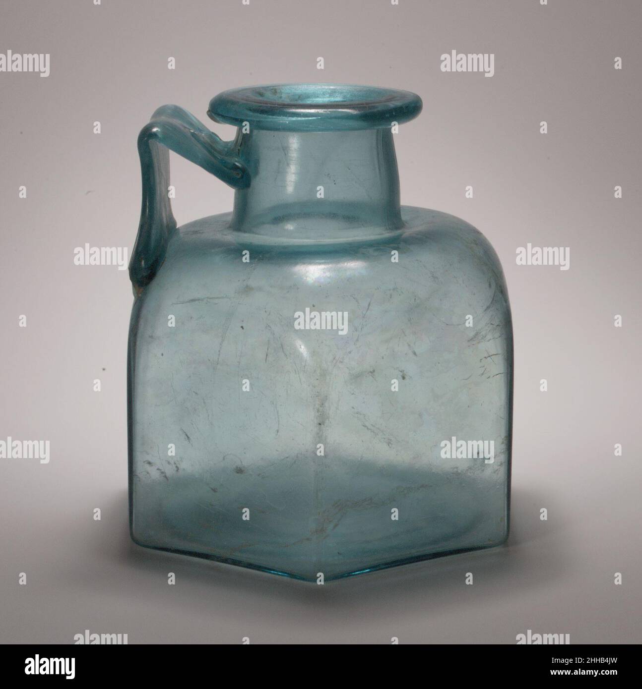 Flacon hexagonal en verre à une poignée fin 1st–début 3rd Century A.D. Roman bleu clair translucide avec teinte verdâtre ; manche de la même couleur. Rebord épais, partiellement tubulaire, plié, vers le bas, rond,et dans ; col cylindrique, évasé vers le bas, avec profil légèrement convexe ; épaulement horizontal, légèrement poussé vers le centre et épaules arrondies inclinées ; corps hexagonal, côtés plats et verticaux ; fond légèrement concave ;main à large bande, épédée sur la surface extérieure, fixée au bord de l'épaule au-dessus d'un côté, tracée verticalement, puis tournée à une poignée aiguë, et traîné sur le cou, avec t Banque D'Images