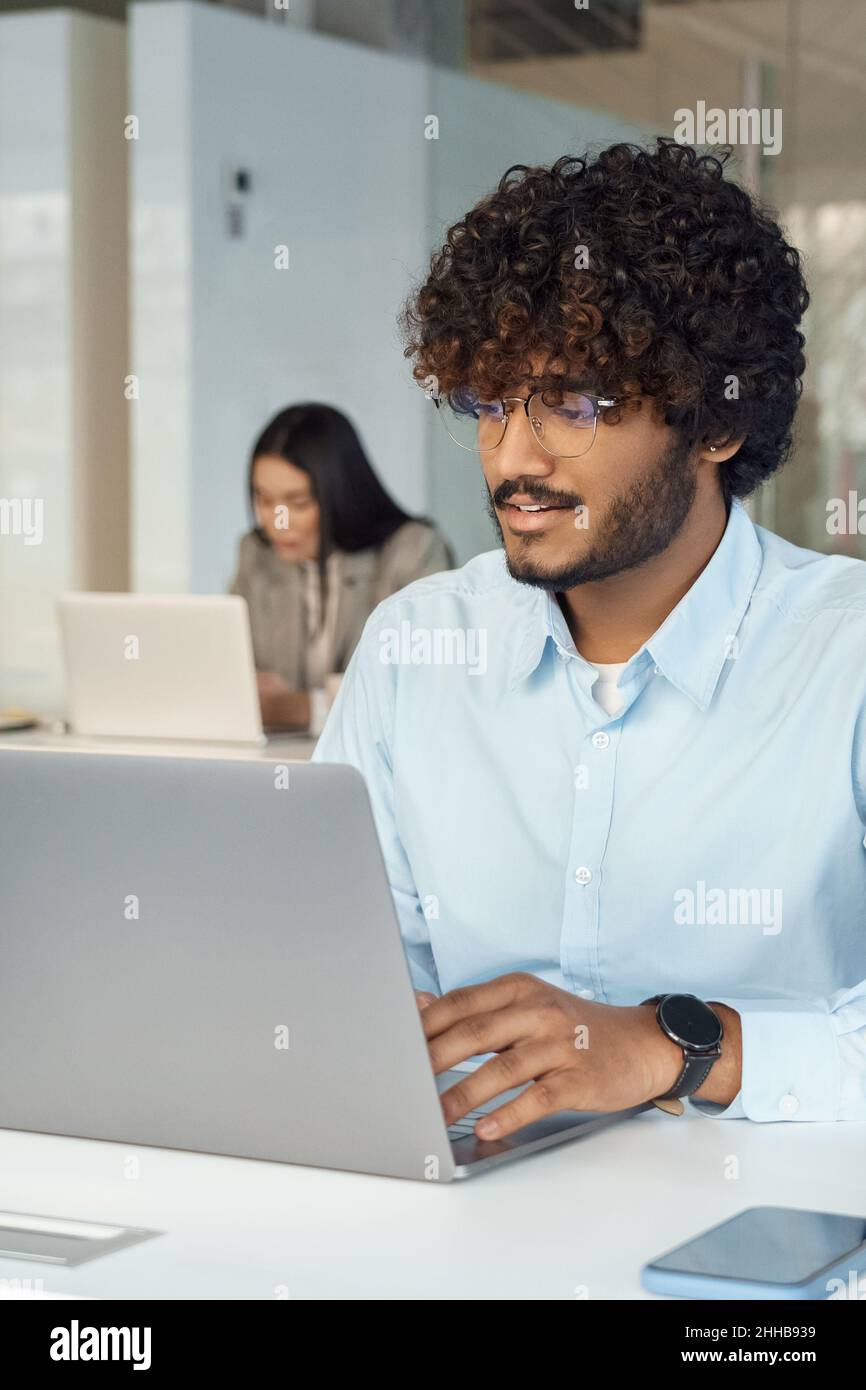 Un jeune indien intelligent et occupé en lunettes travaille sur un ordinateur portable, assis au bureau Banque D'Images