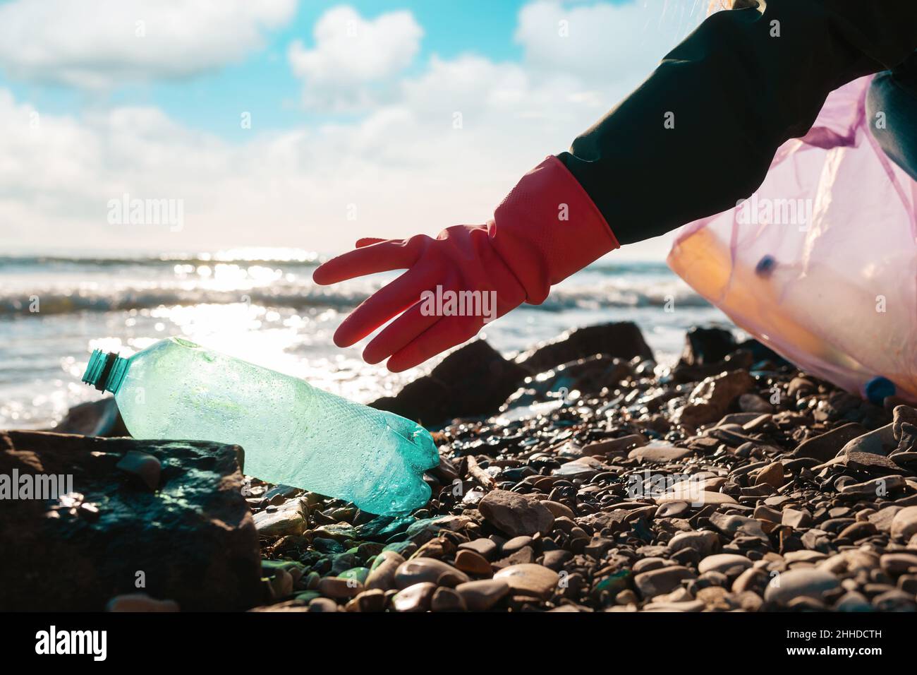 Un volontaire en gants de caoutchouc atteint pour une bouteille verte en plastique sale qui se trouve sur la rive de l'océan.Gros plan à la main.Concept de Save the Planet. Banque D'Images