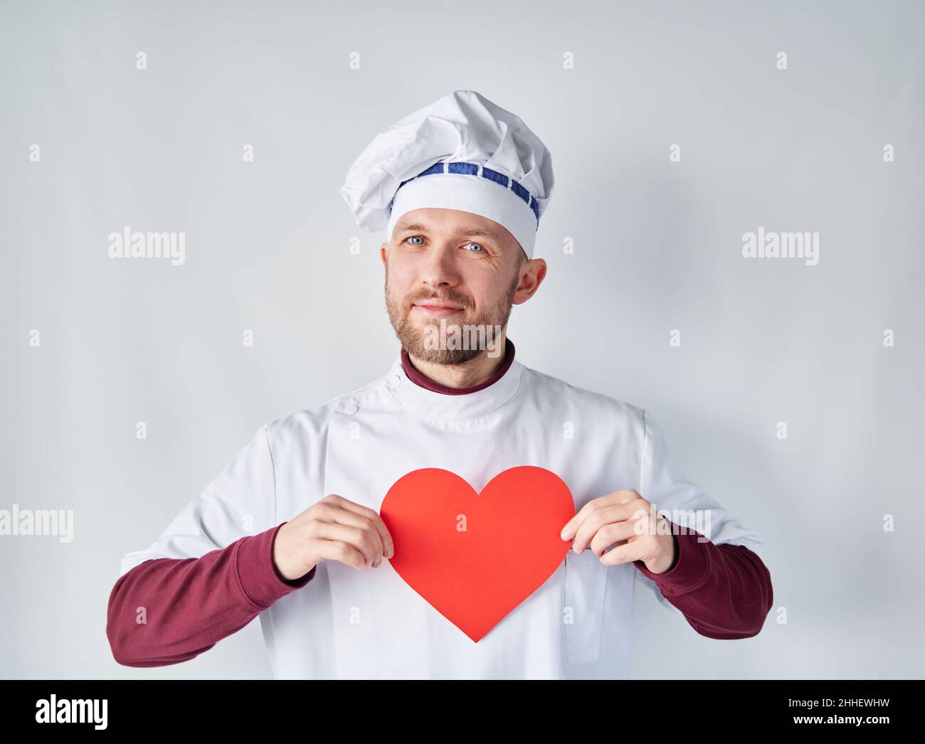 Chef attrayant tenant un coeur rouge devant le vêtement de travail.Chef barbu ou boulanger en uniforme blanc avec coeur en papier rouge en forme de Saint-Valentin, romance ou amour concept.Photo de haute qualité Banque D'Images